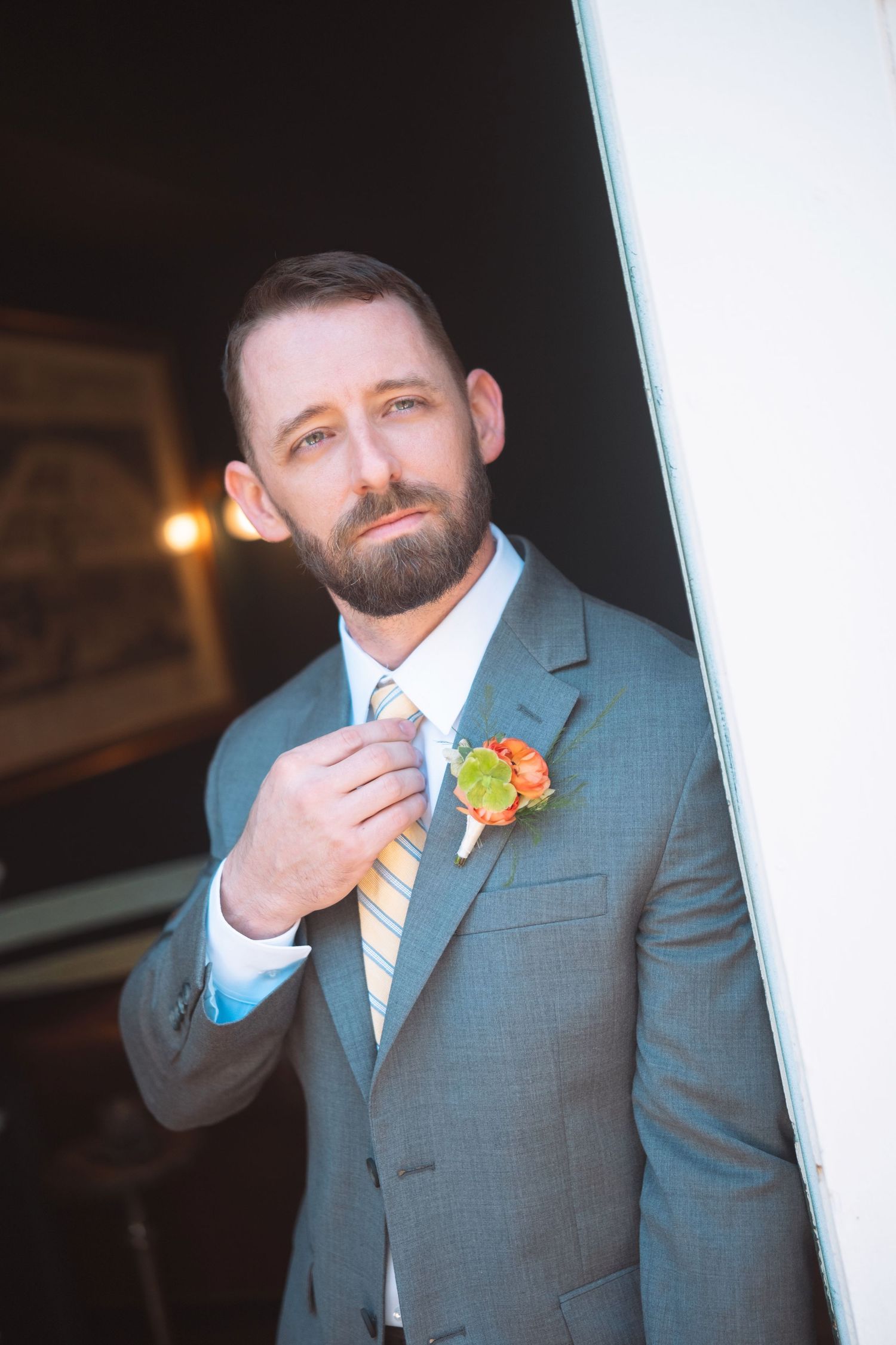 Dapper groom in gray suit adjusting his yellow tie with an orange boutonniere.