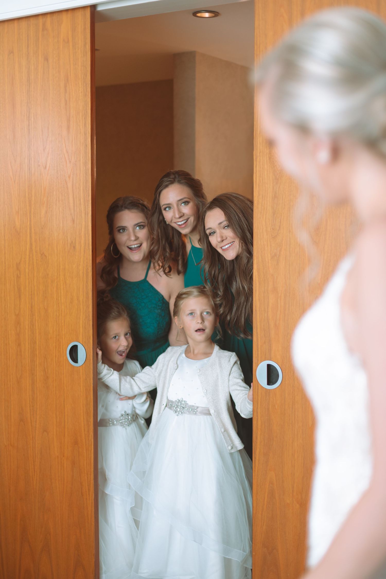 Bridesmaids and flower girls peek through wooden doors at bride in white wedding dress.