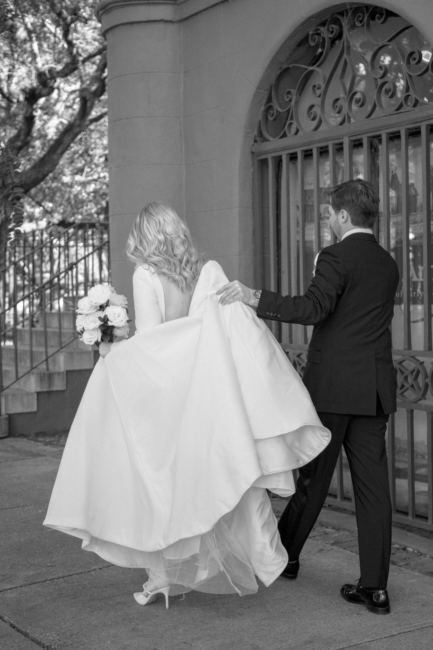 Black and white photo of bride and groom walking through ornate iron gate at wedding venue.