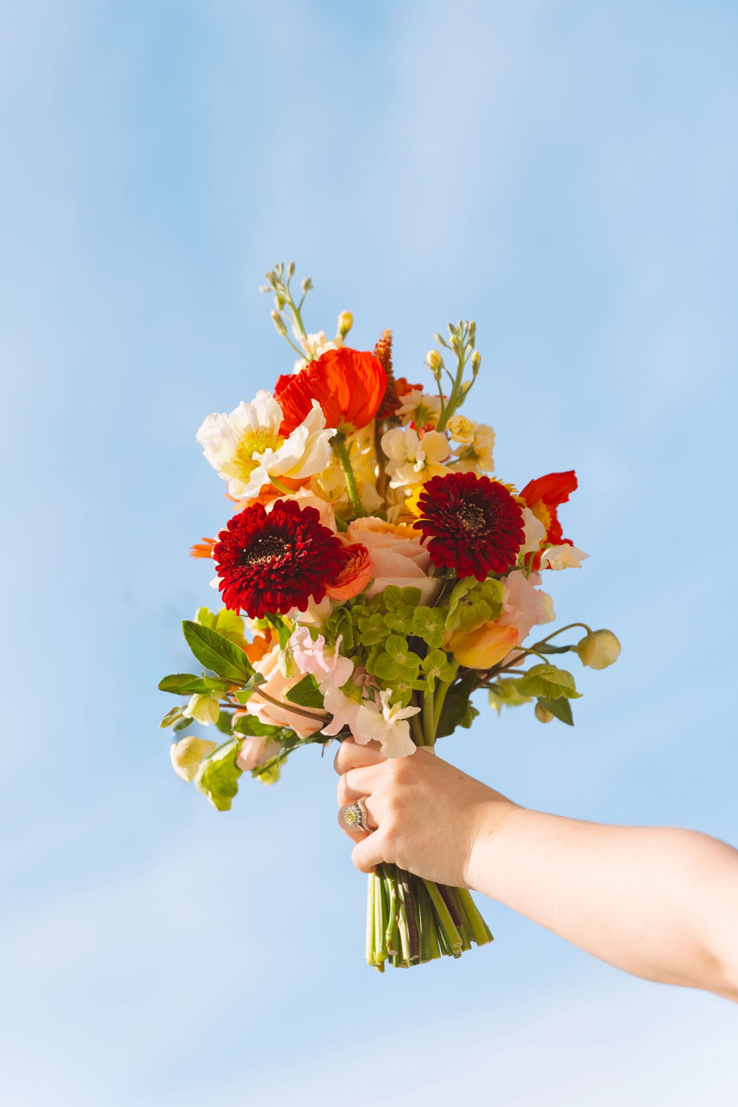 Hand holding a vibrant bridal bouquet of red and orange flowers against a blue sky.