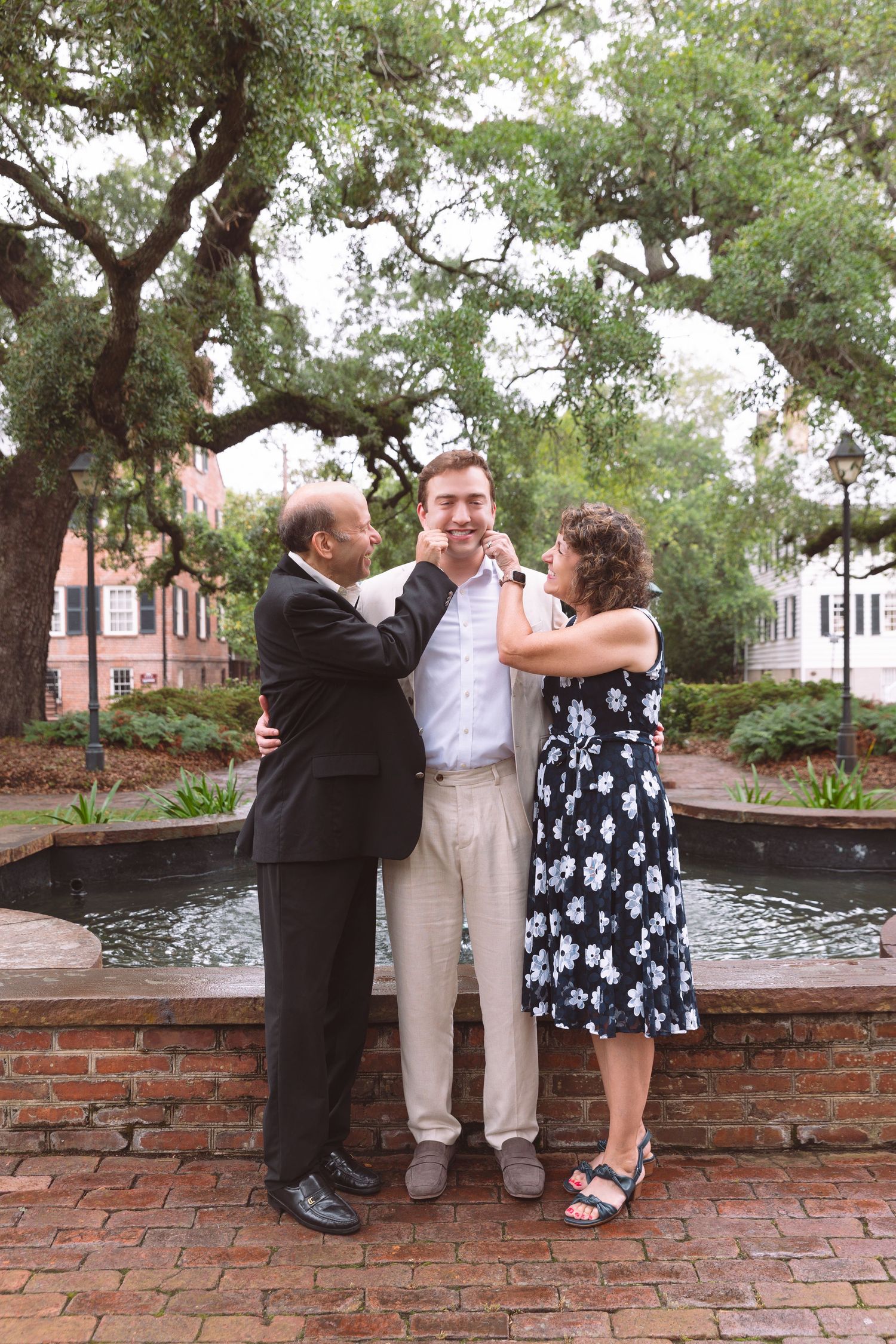 Family portrait of parents proudly adjusting their son's collar near a brick fountain in a lush garden.