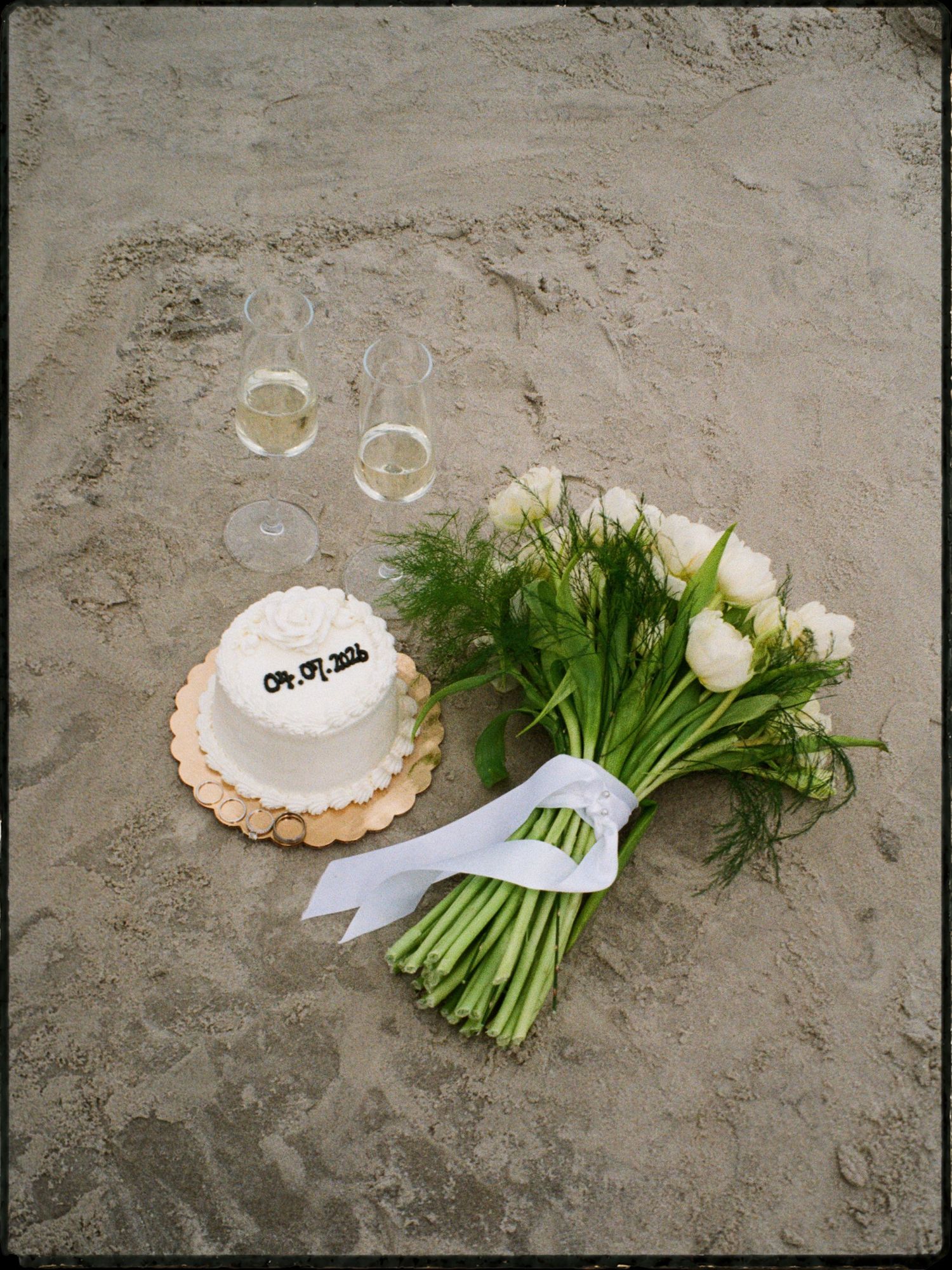 Wedding flat lay on sandy beach featuring small cake, white rose bouquet, and champagne glasses.