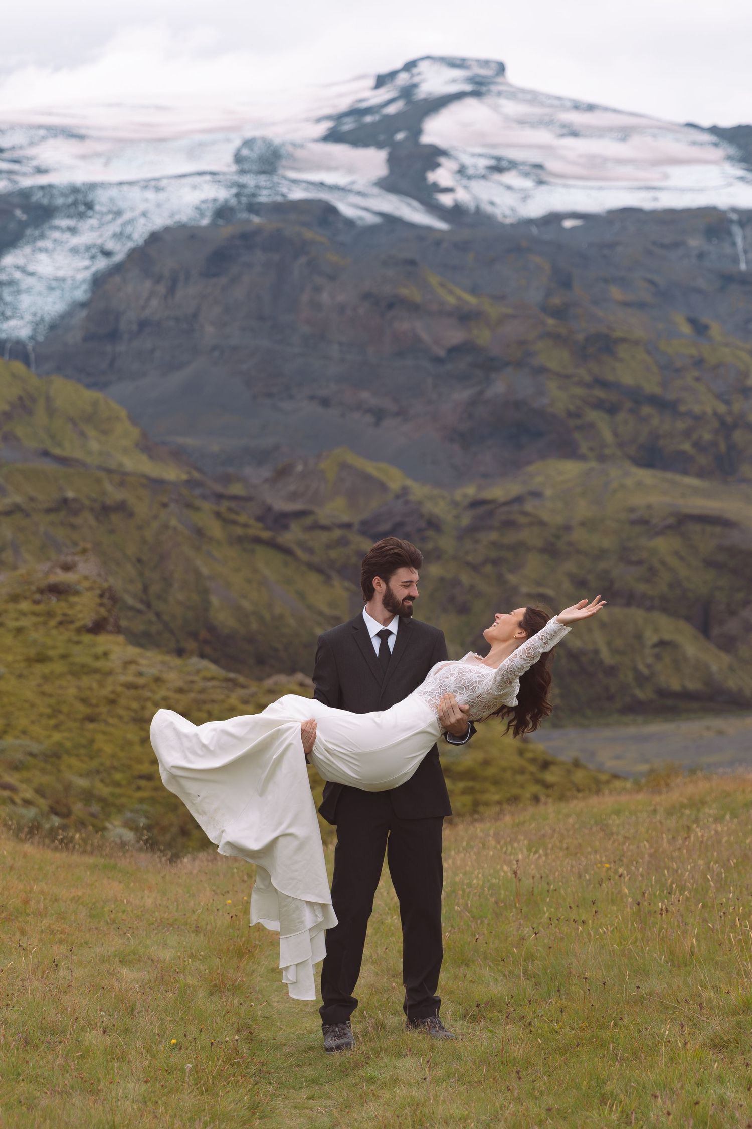Groom carries bride in his arms as she leans back joyfully in a playful outdoor mountain wedding photo.