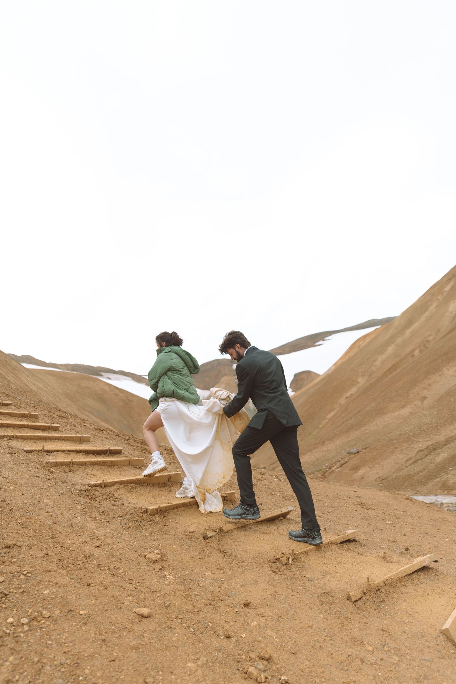 Groom carries bride's white dress train while climbing wooden steps on Iceland's volcanic mountain slope.