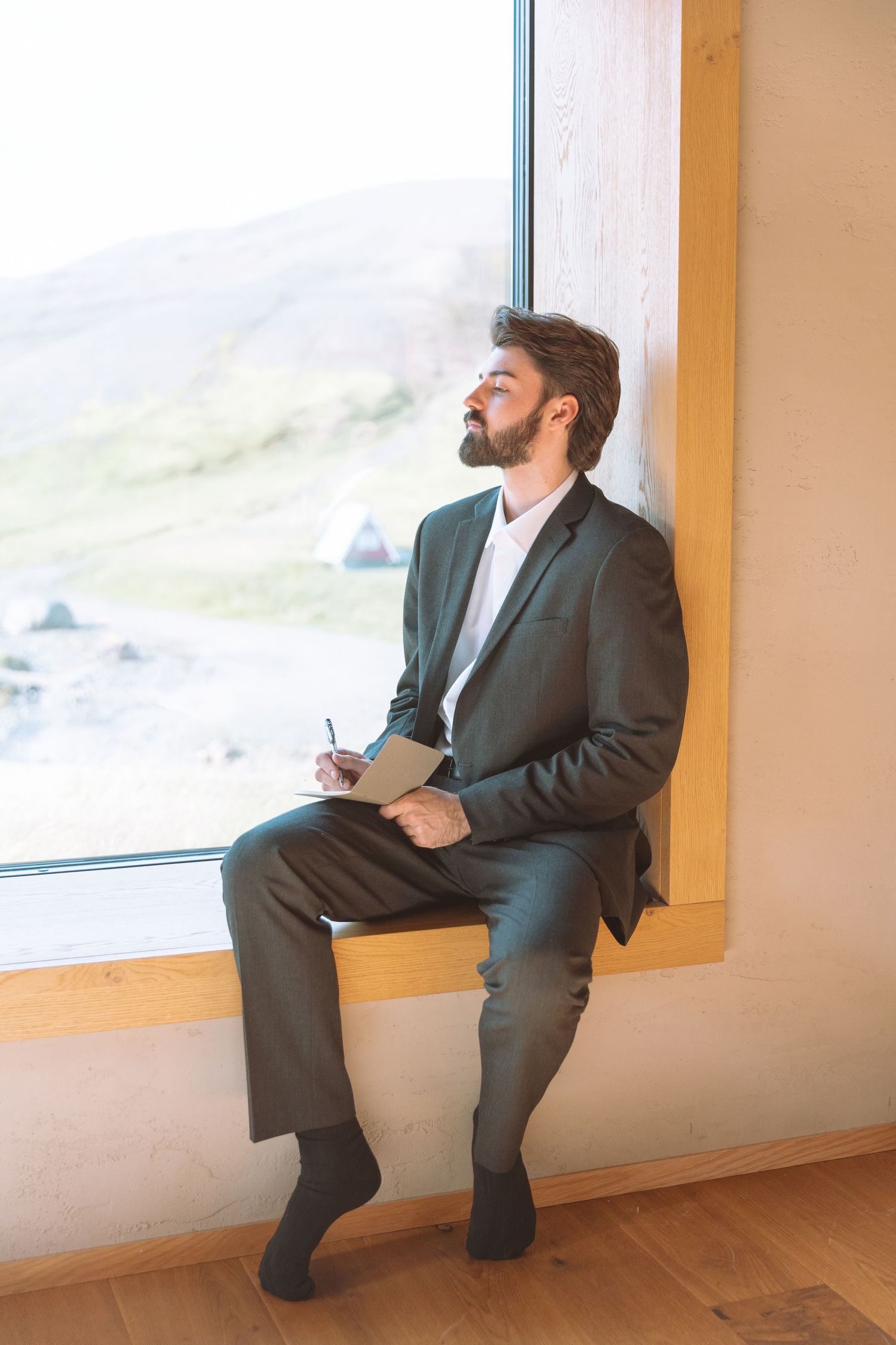 Bearded groom in dark suit sits on window ledge writing vows in notebook with mountain views.