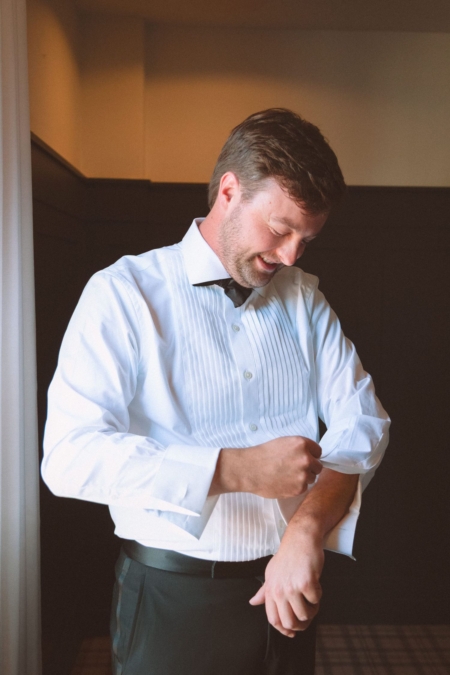 Smiling groom in white tuxedo shirt fastening cufflinks while getting ready for wedding day.