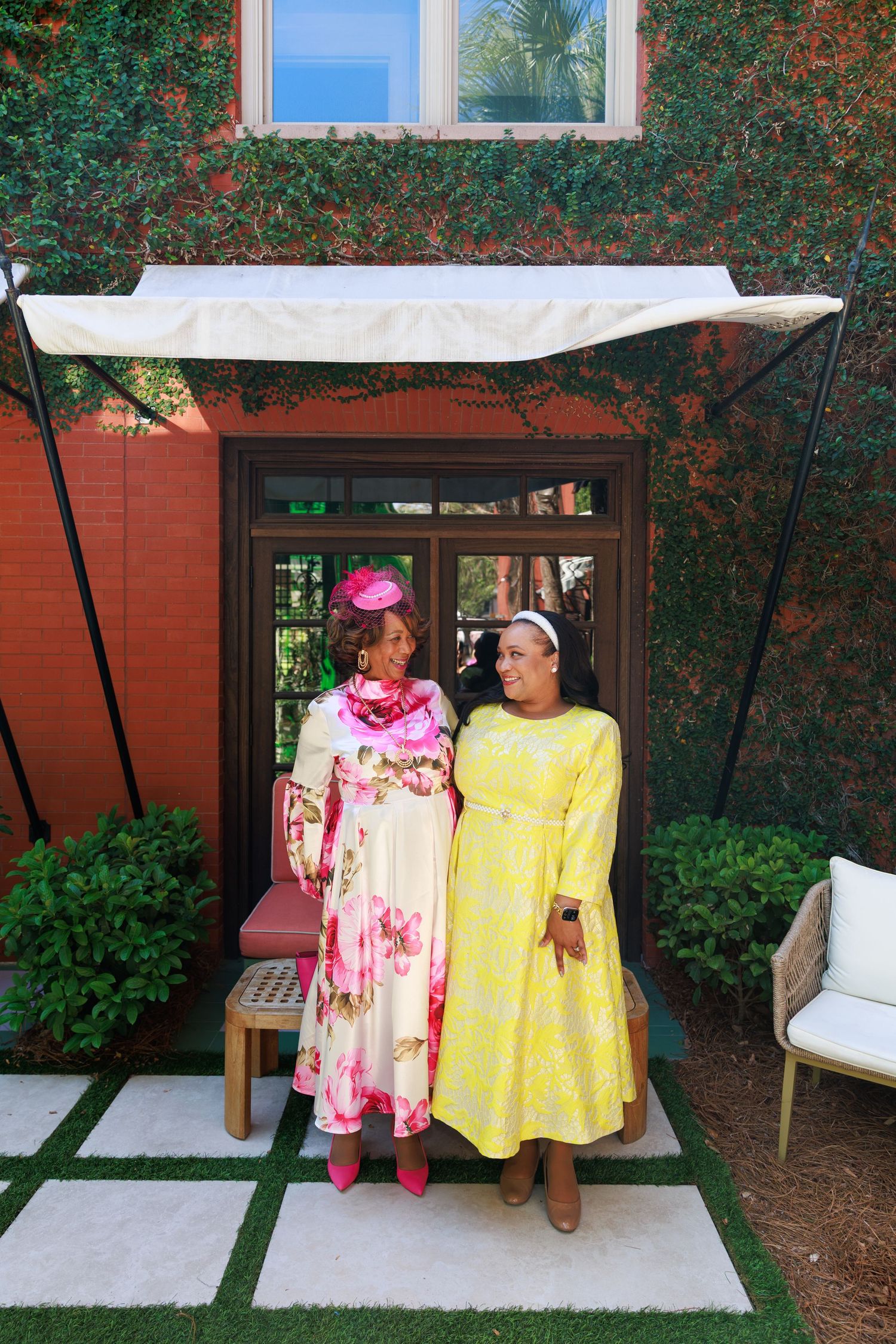 Two women in colorful floral dresses smiling together outside a brick building with lush greenery.