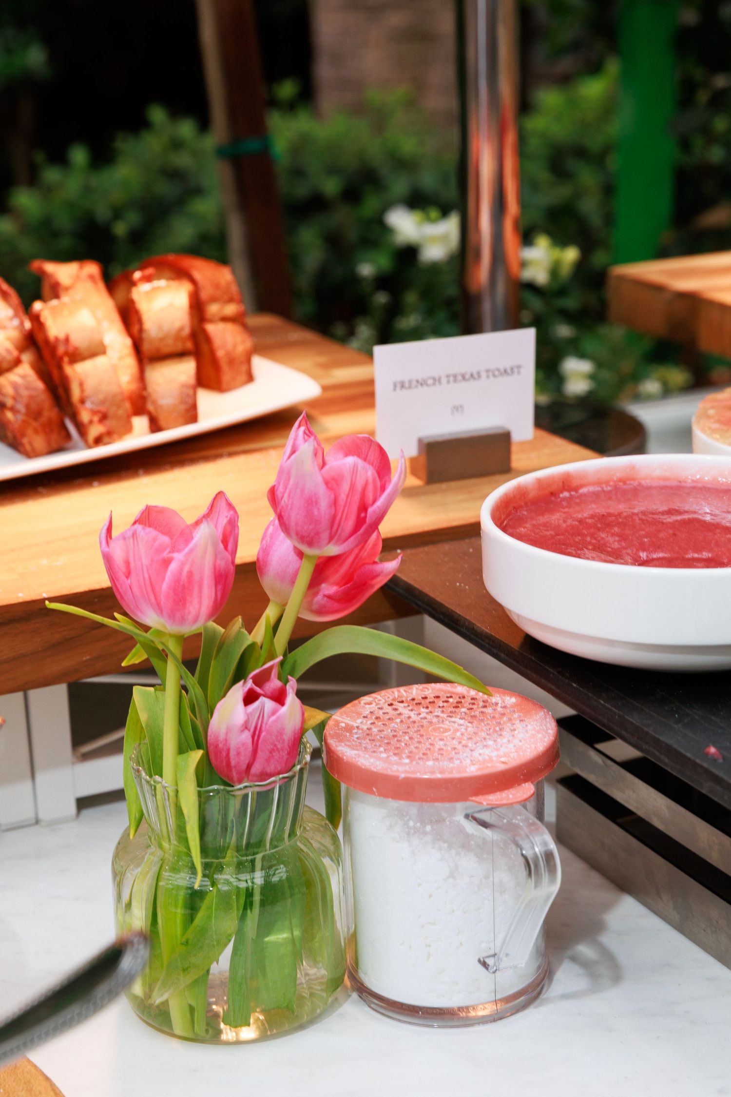 Pink tulips in glass vase beside French toast and strawberry soup on a wooden breakfast buffet table.
