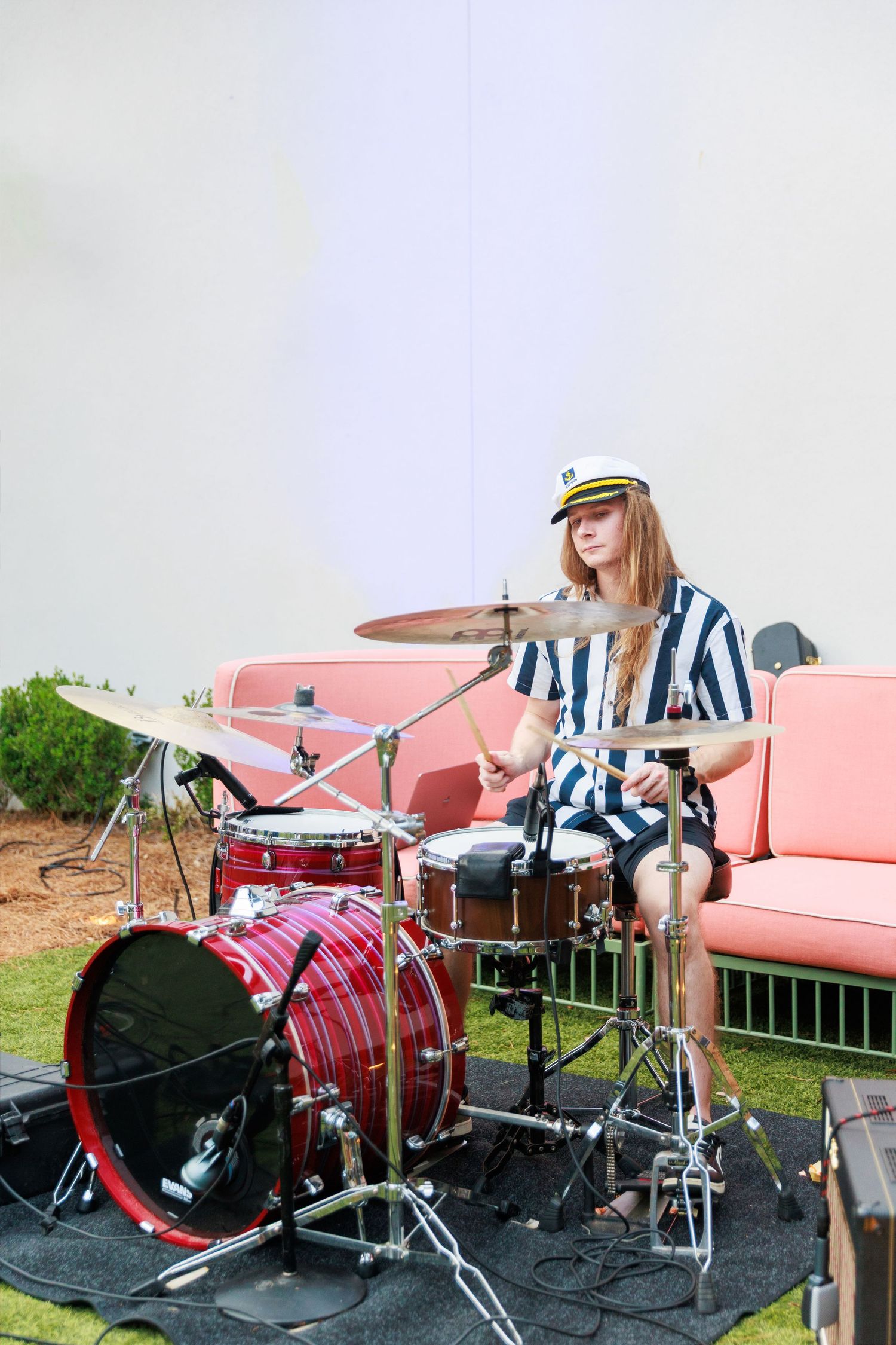 Young woman in striped outfit and captain hat playing drums outdoors at a live music event.
