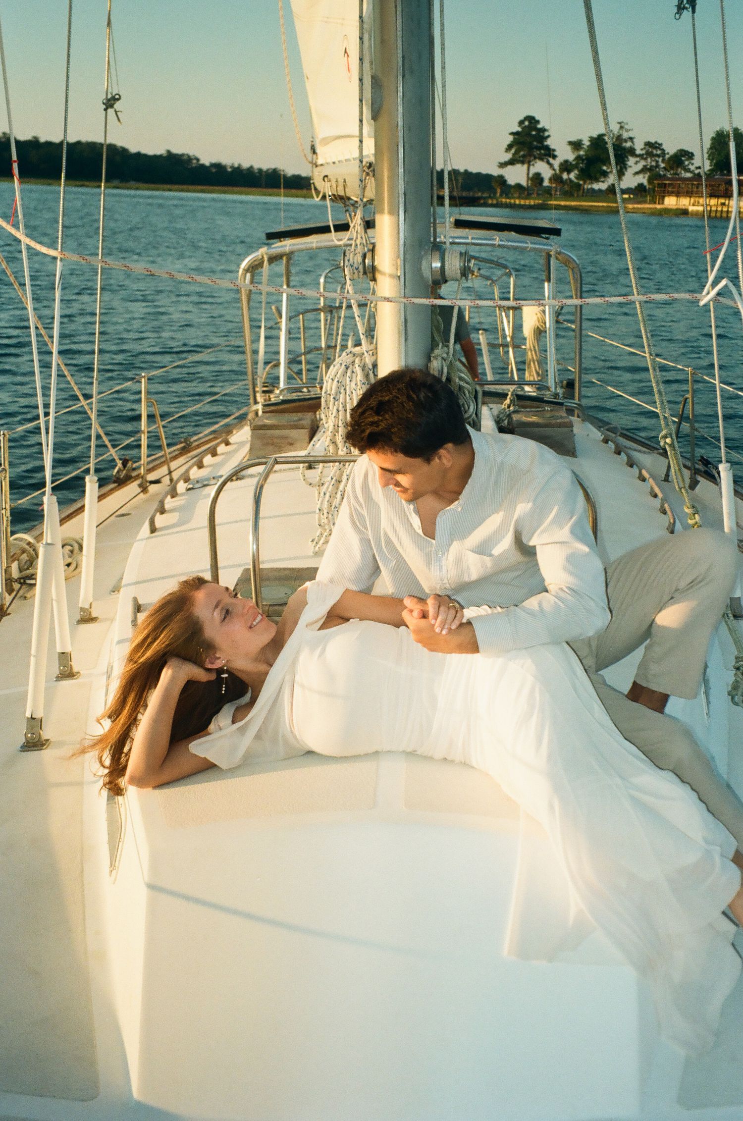 Romantic couple in white outfits relaxing on a sailboat deck with calm water and trees in the background.
