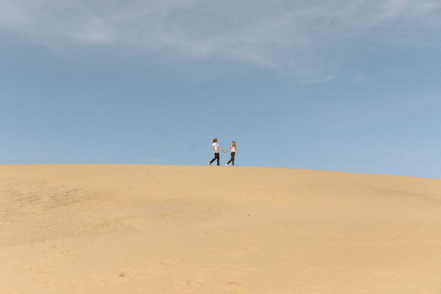 Two people walking along the ridge of a vast sandy desert dune under a clear blue sky.