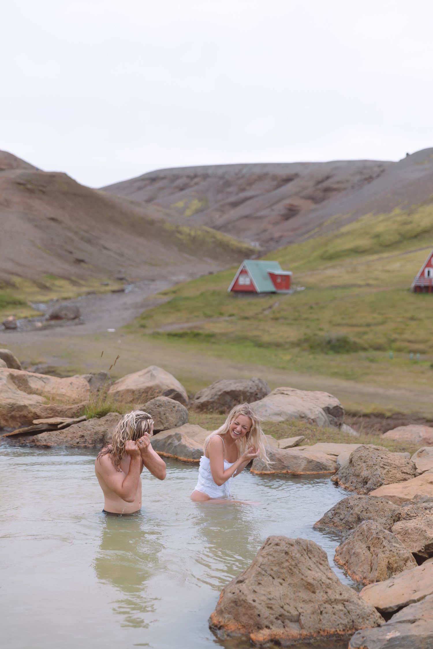 Two women relaxing in a natural hot spring pool with colorful mountain cabins in background.