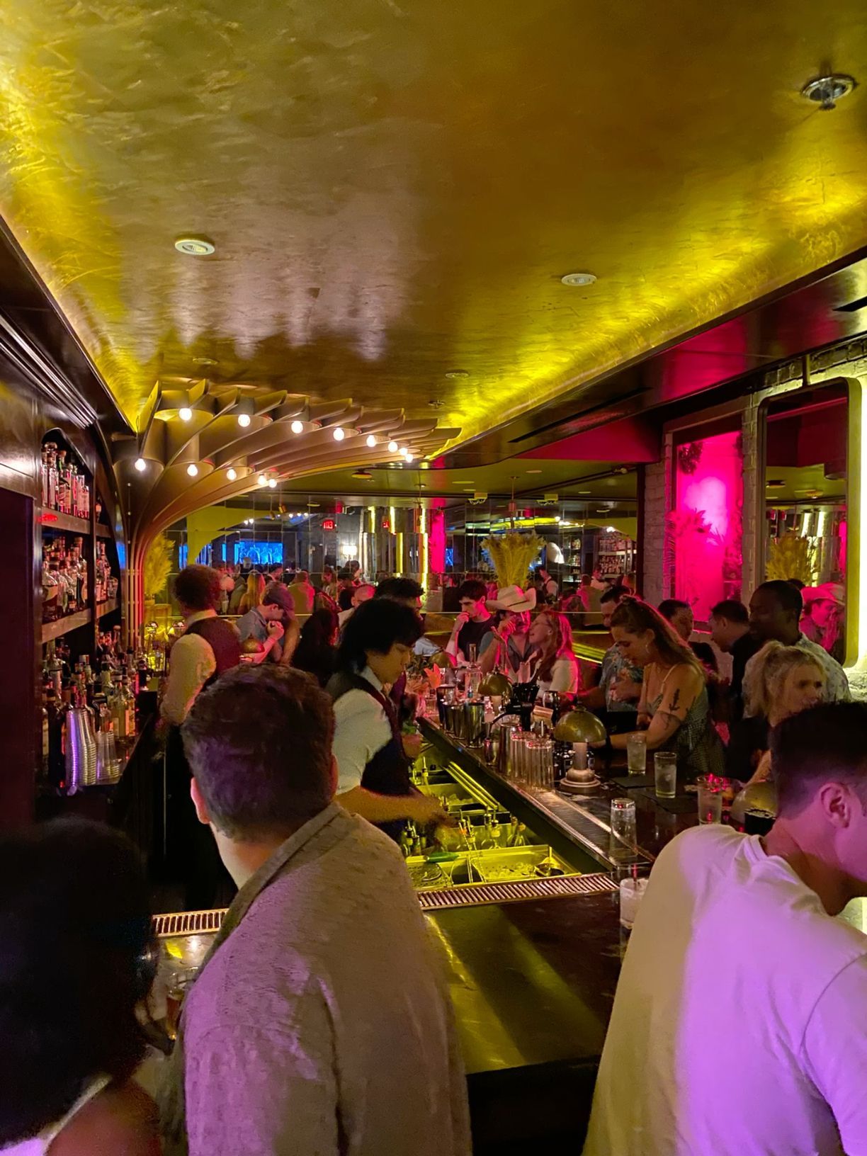 Lively crowded bar with gold ceiling, neon pink lights, and busy bartenders serving drinks.