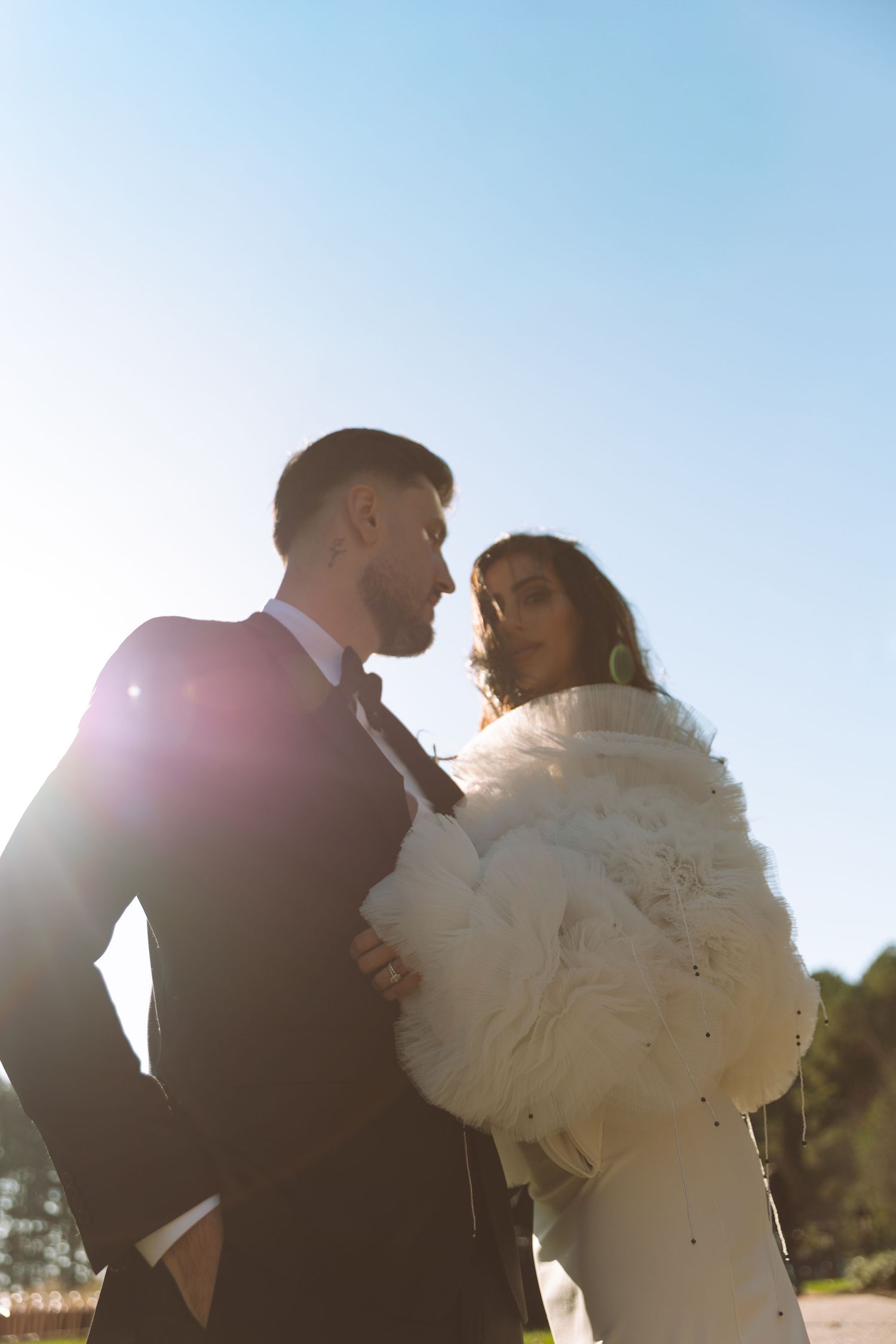 Bride in white fur coat and groom embracing outdoors in bright sunlight.