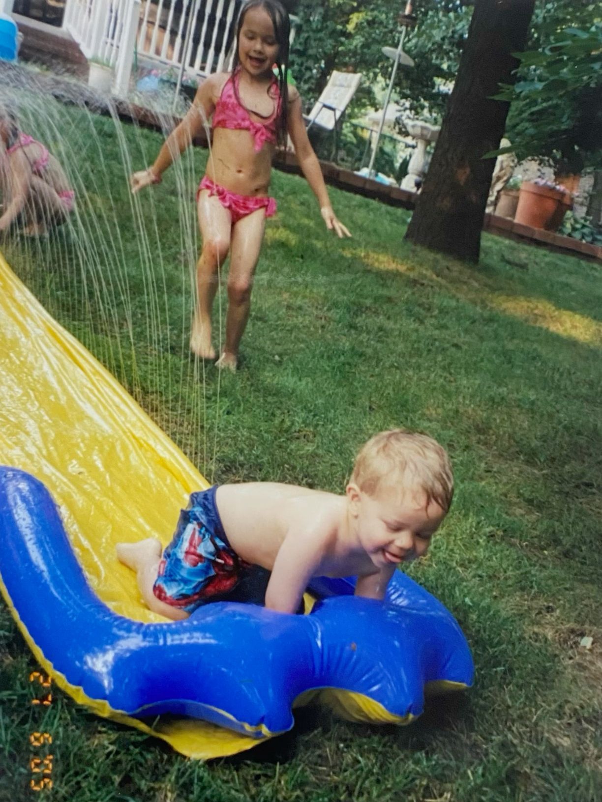 Two children playing on a backyard slip and slide on a sunny summer day, one sliding down while another waits in a pink bikini.