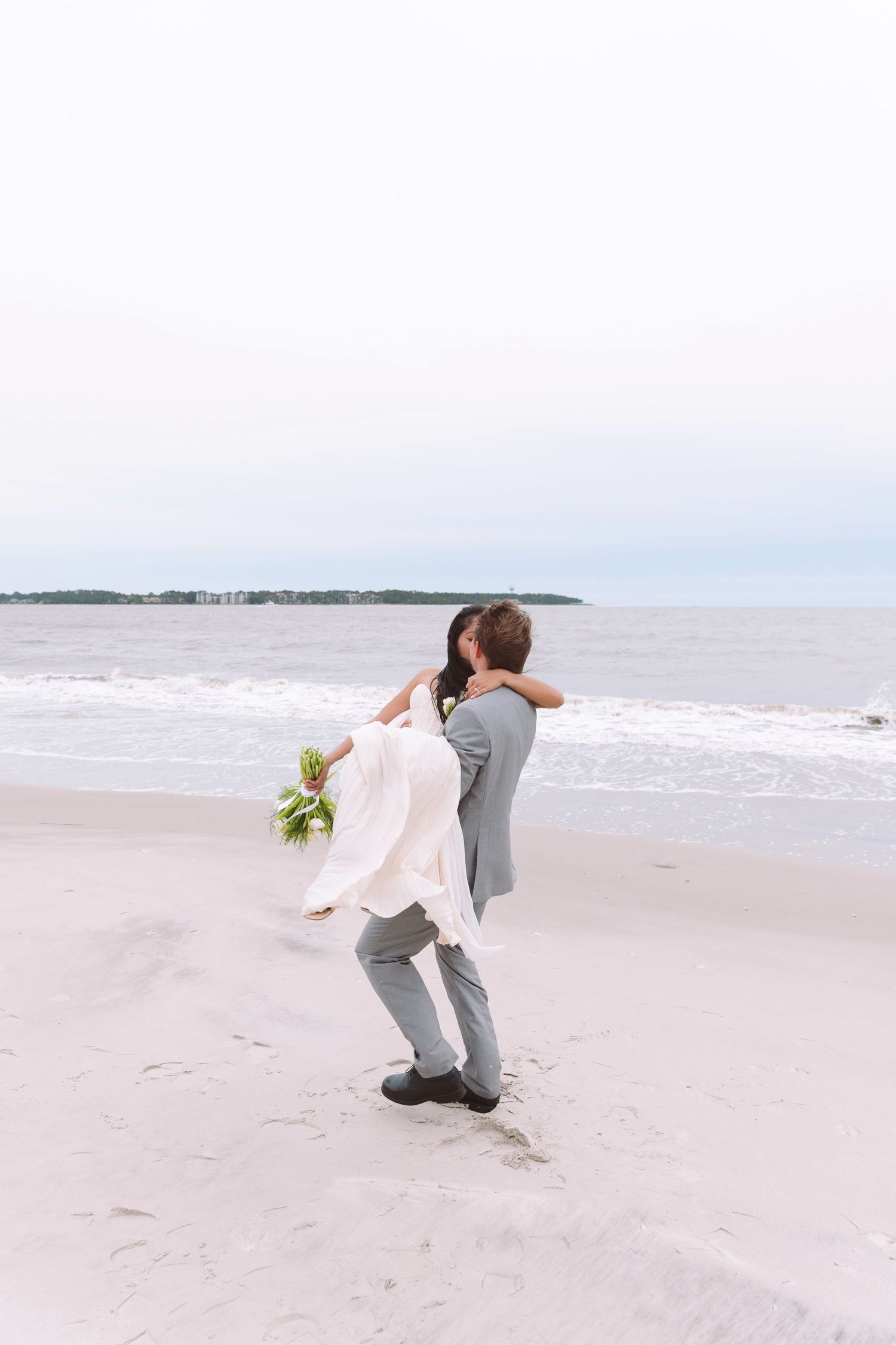 Groom in gray suit lifts bride in white gown on sandy beach with ocean waves behind them.
