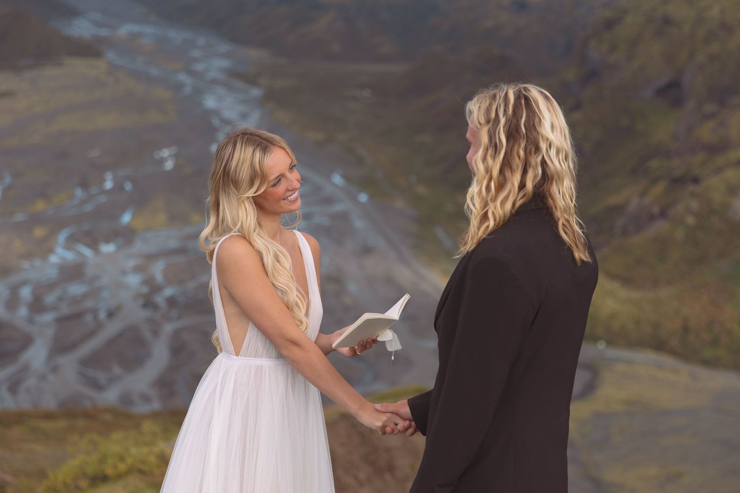 Two brides holding hands during outdoor Iceland elopement ceremony with dramatic river valley backdrop.