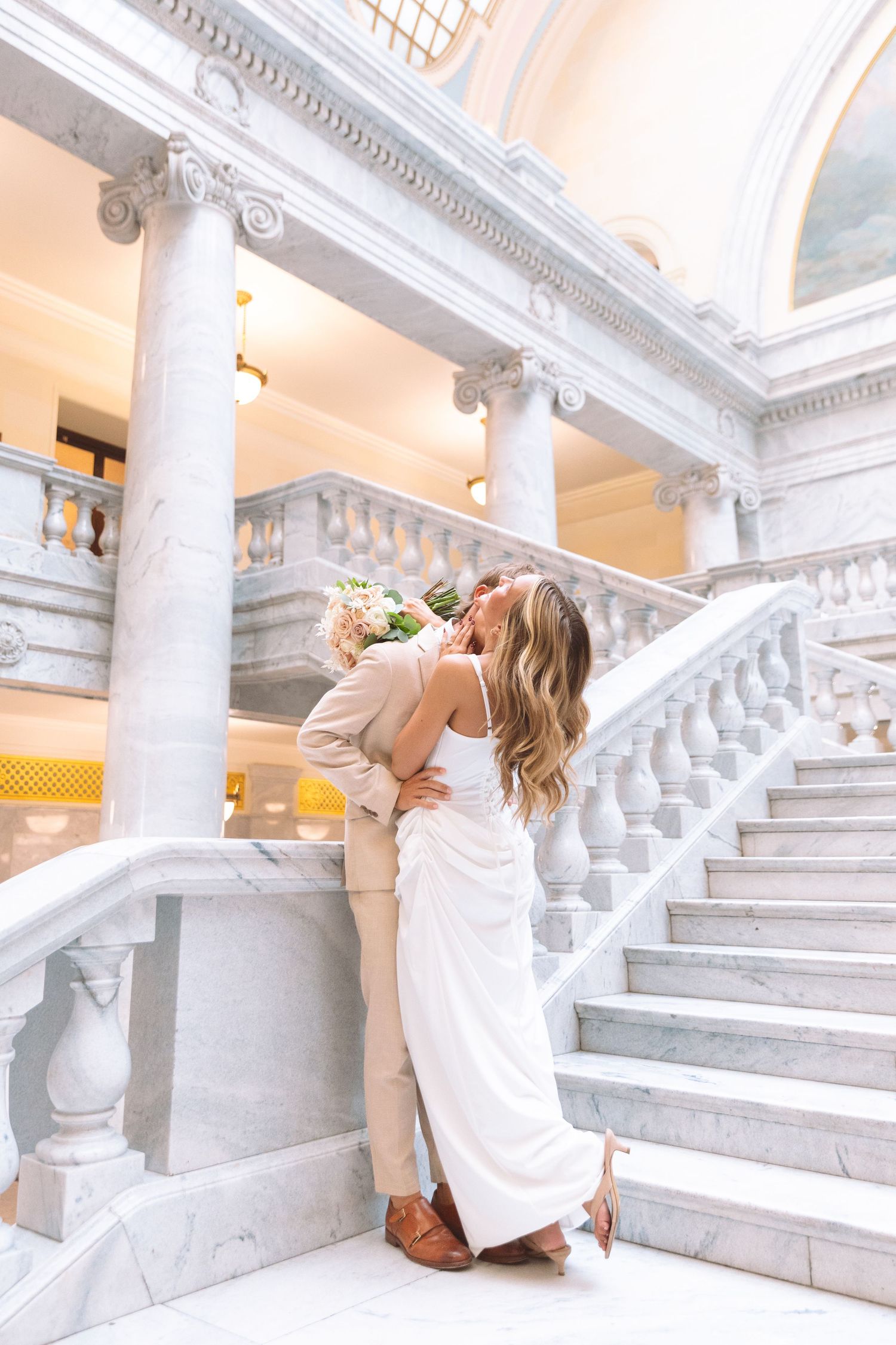 Bride and groom share passionate kiss on marble staircase inside elegant white stone building.
