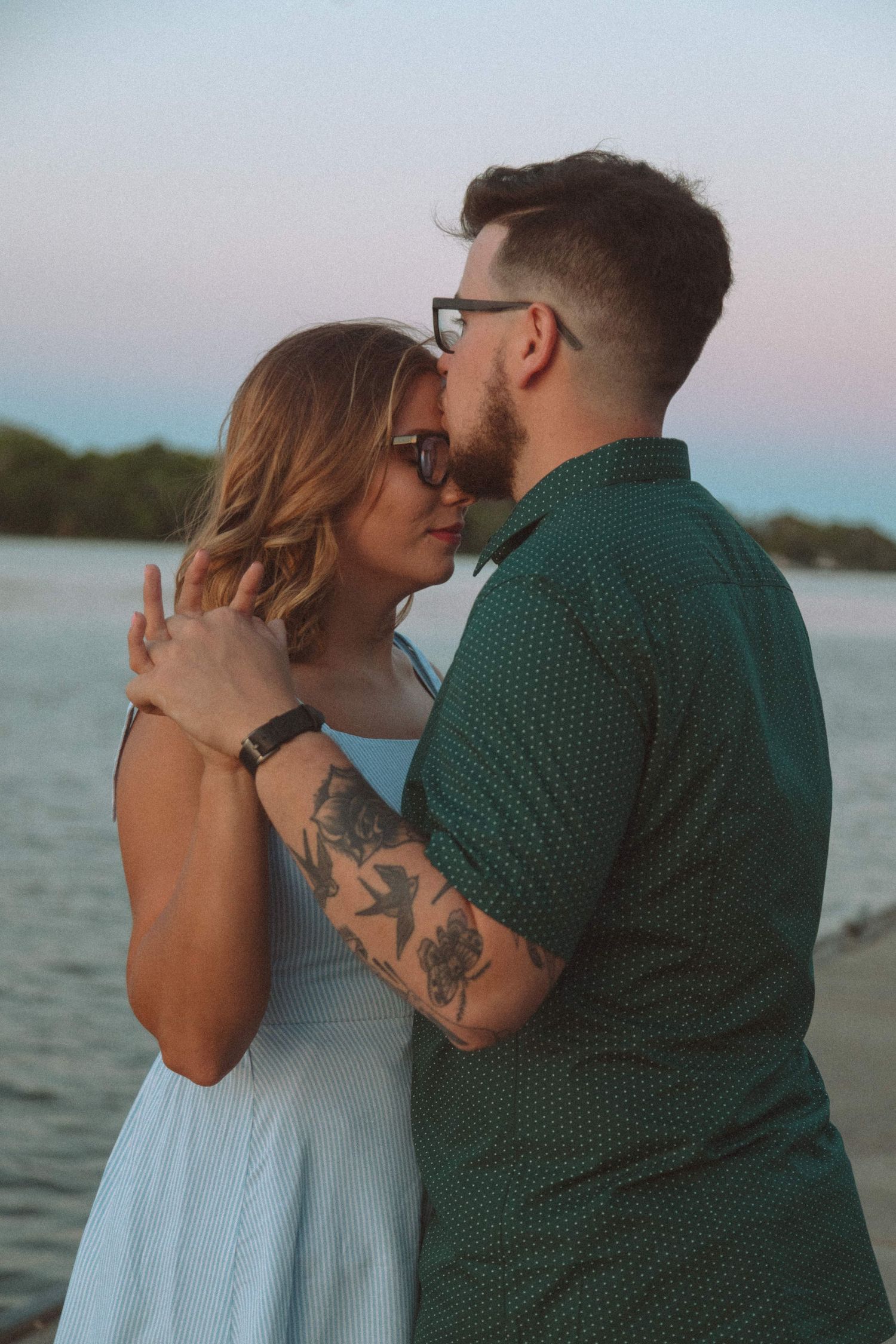 Couple sharing a tender moment near a lake at sunset, man with tattoos and glasses embracing woman in white dress.