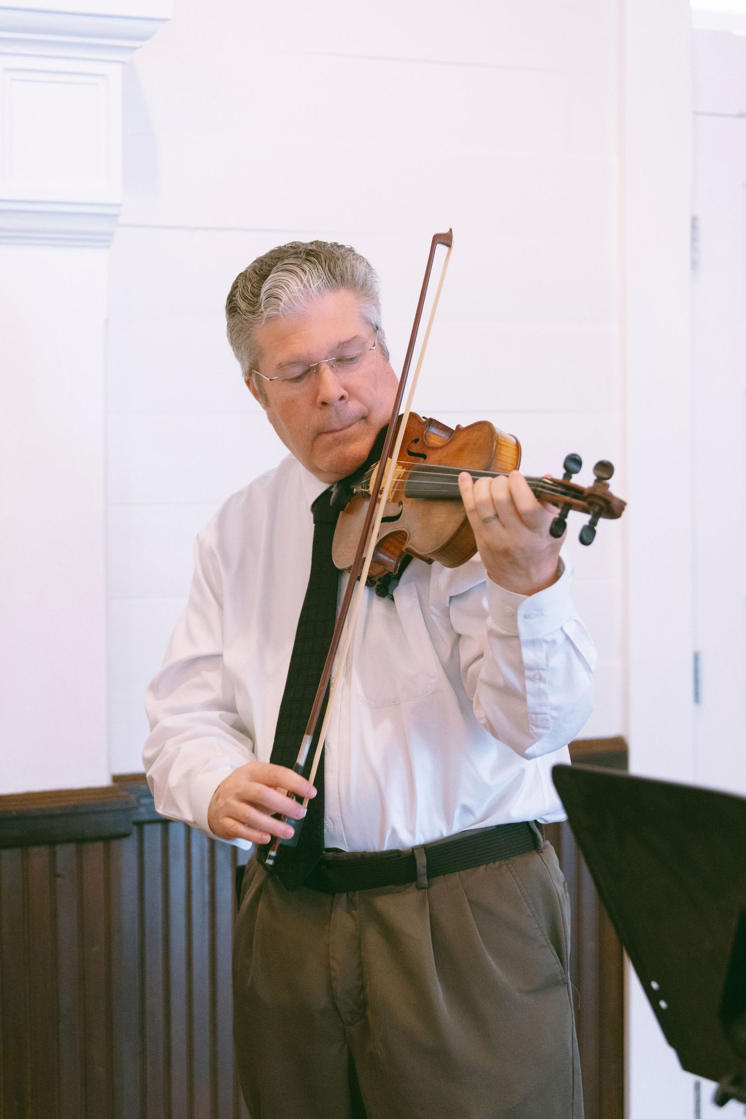 Middle-aged man playing violin in white shirt and tie, performing in a bright indoor setting.