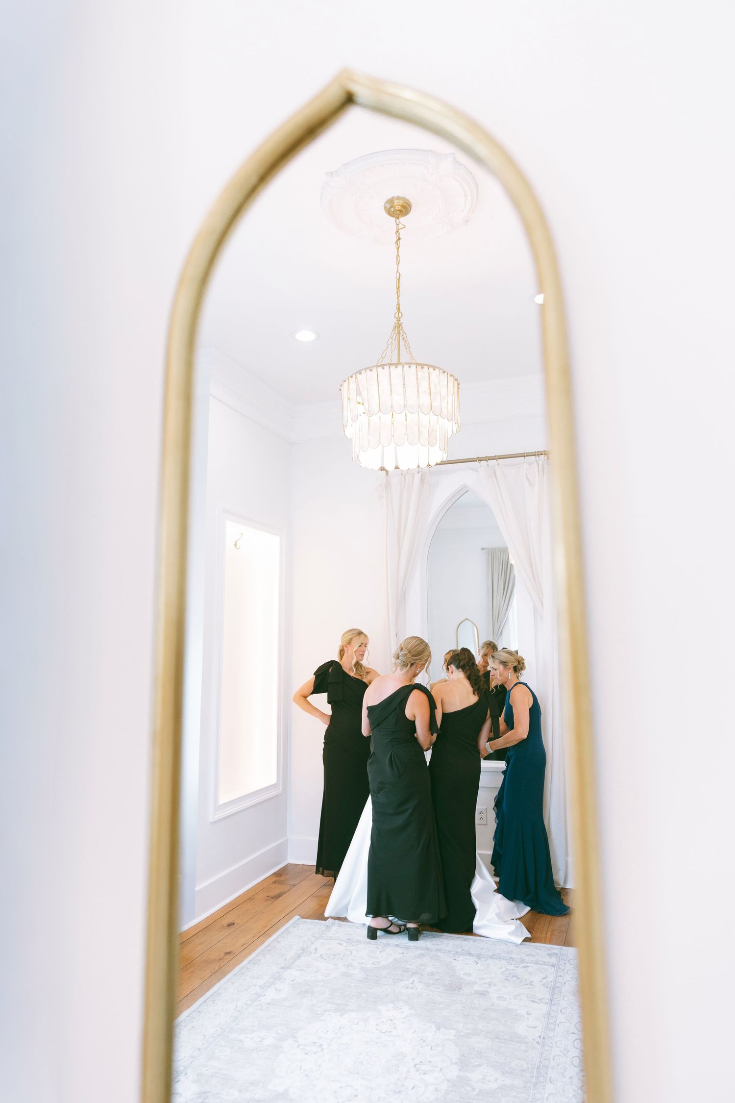 Bridesmaids in black gowns gather around bride reflected in gold arch mirror in bright white bridal suite.