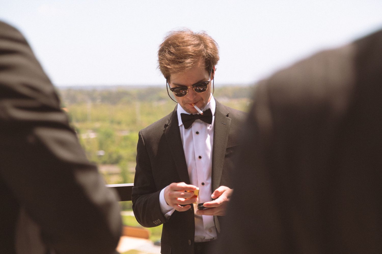 Dapper groom in black tuxedo and bow tie checks his phone while wearing sunglasses before the wedding ceremony.