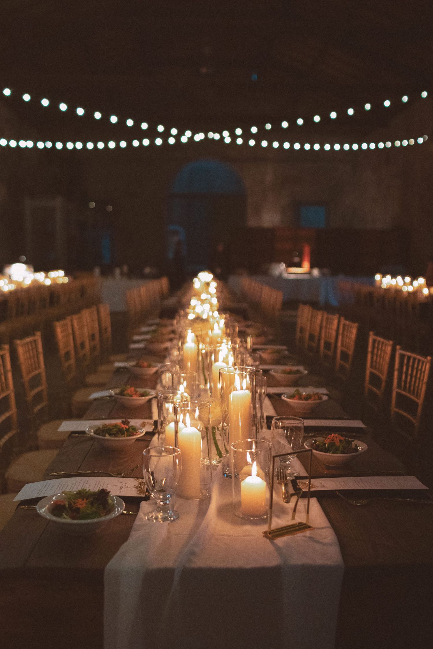 Romantic candlelit wedding reception with long banquet tables, white candles, and bistro lights in a dark hall.