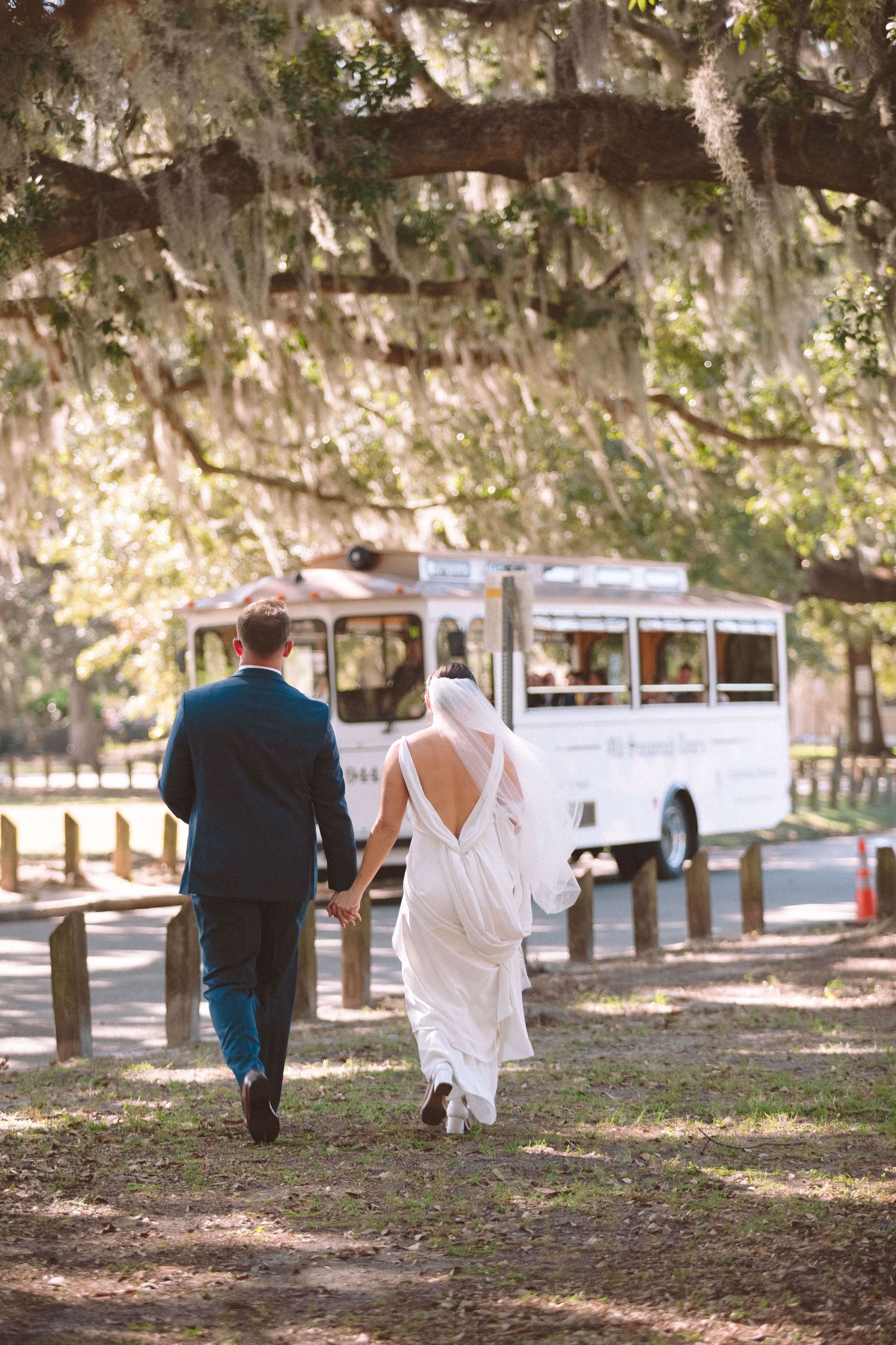 Bride and groom walk hand-in-hand toward vintage white bus under Spanish moss-draped oak trees.