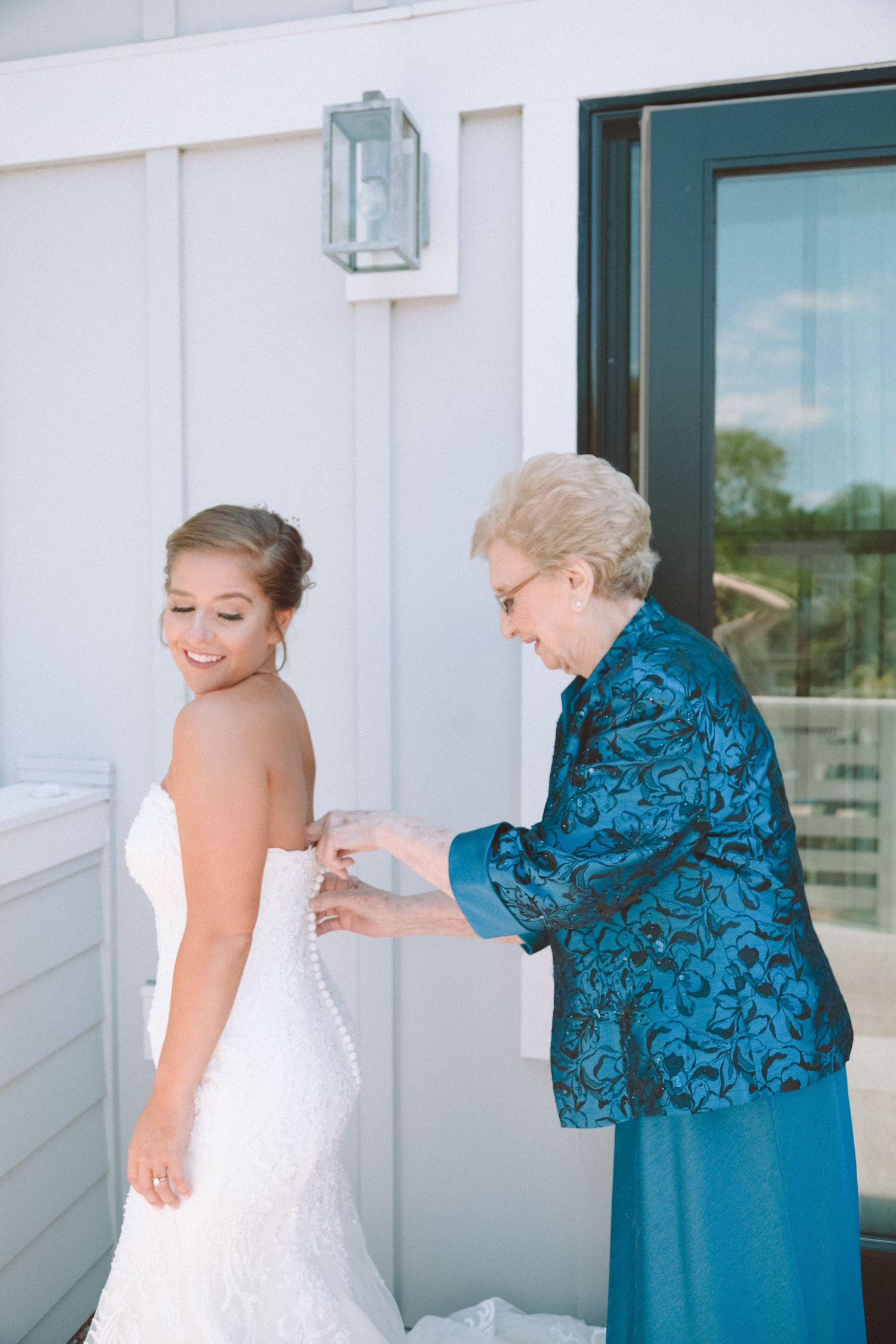 Elderly woman helping a smiling bride button her white lace wedding gown on a porch.