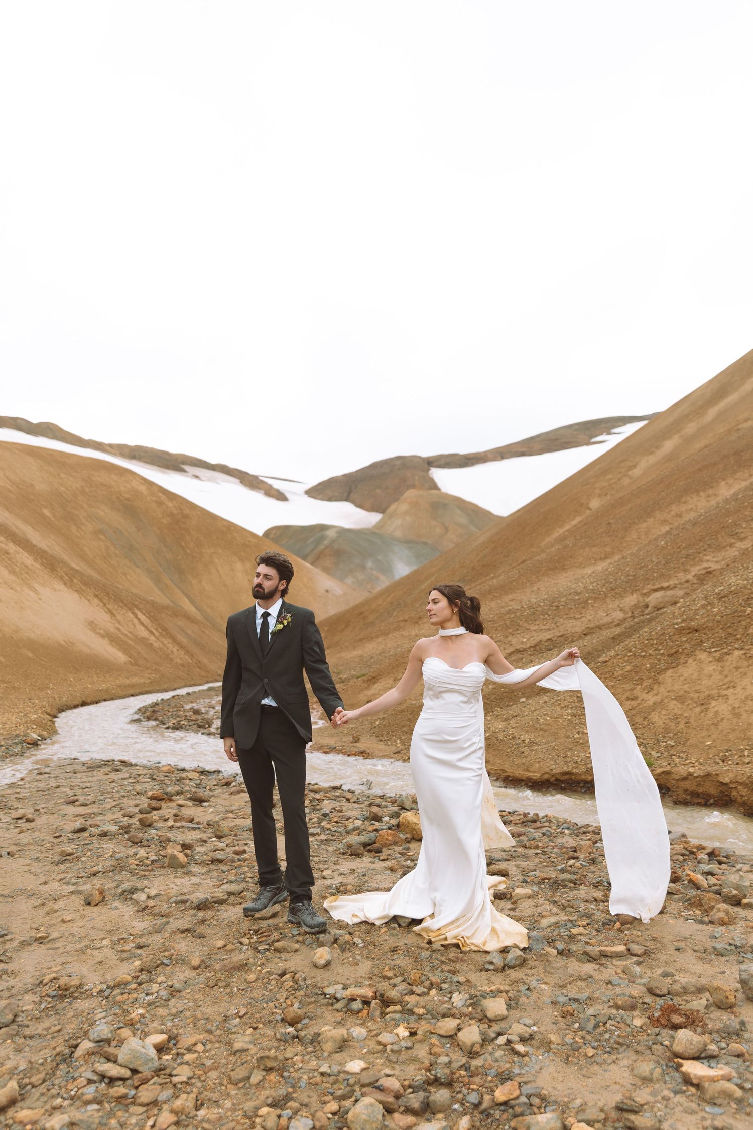 Bride in flowing white dress holds groom's hand while her veil blows in wind against mountain backdrop.