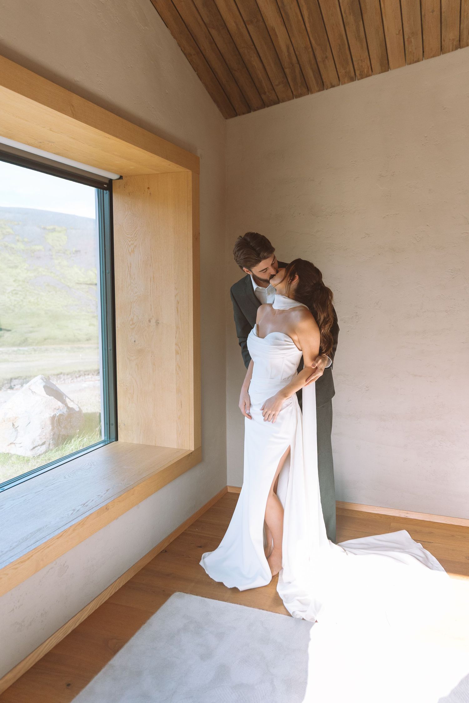 Bride and groom sharing a romantic embrace in a modern minimalist room with large window views.
