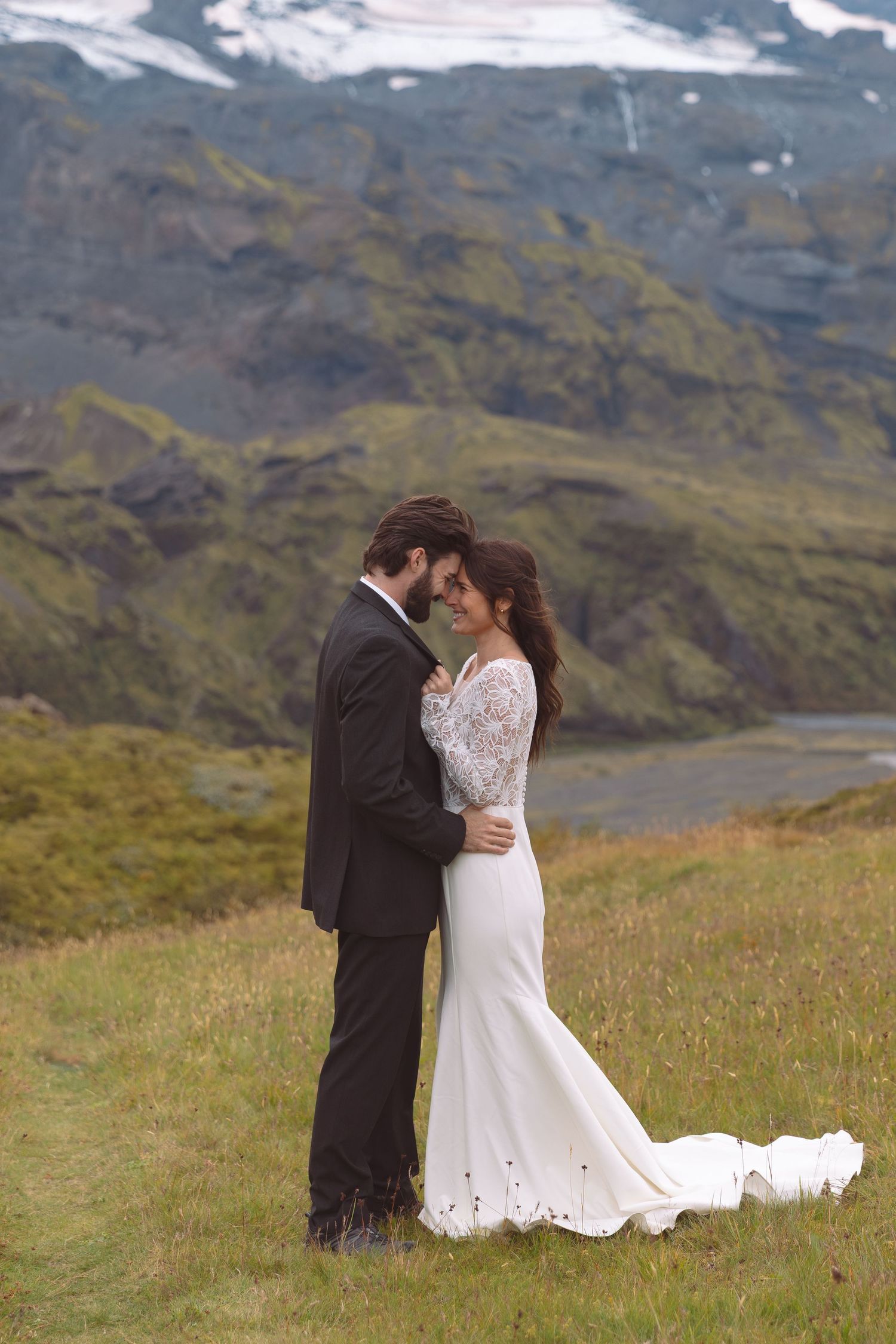 Bride in lace gown and groom in dark suit embracing romantically with Icelandic glacier mountains backdrop.