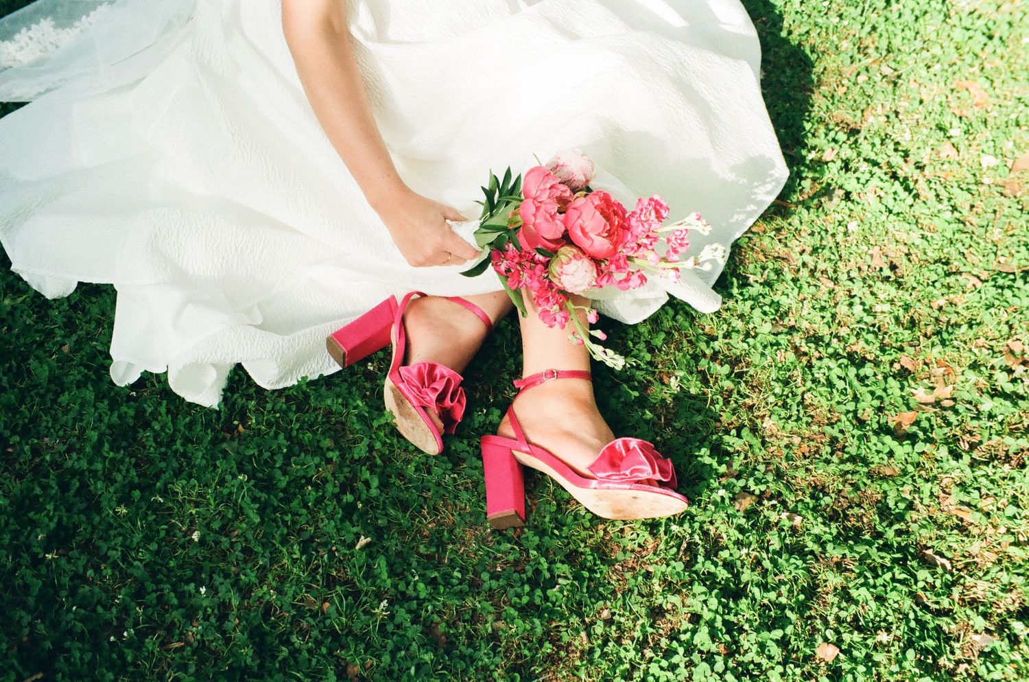 Bride in white gown sitting on green grass with pink peony bouquet and matching pink block heel sandals with bow detail.