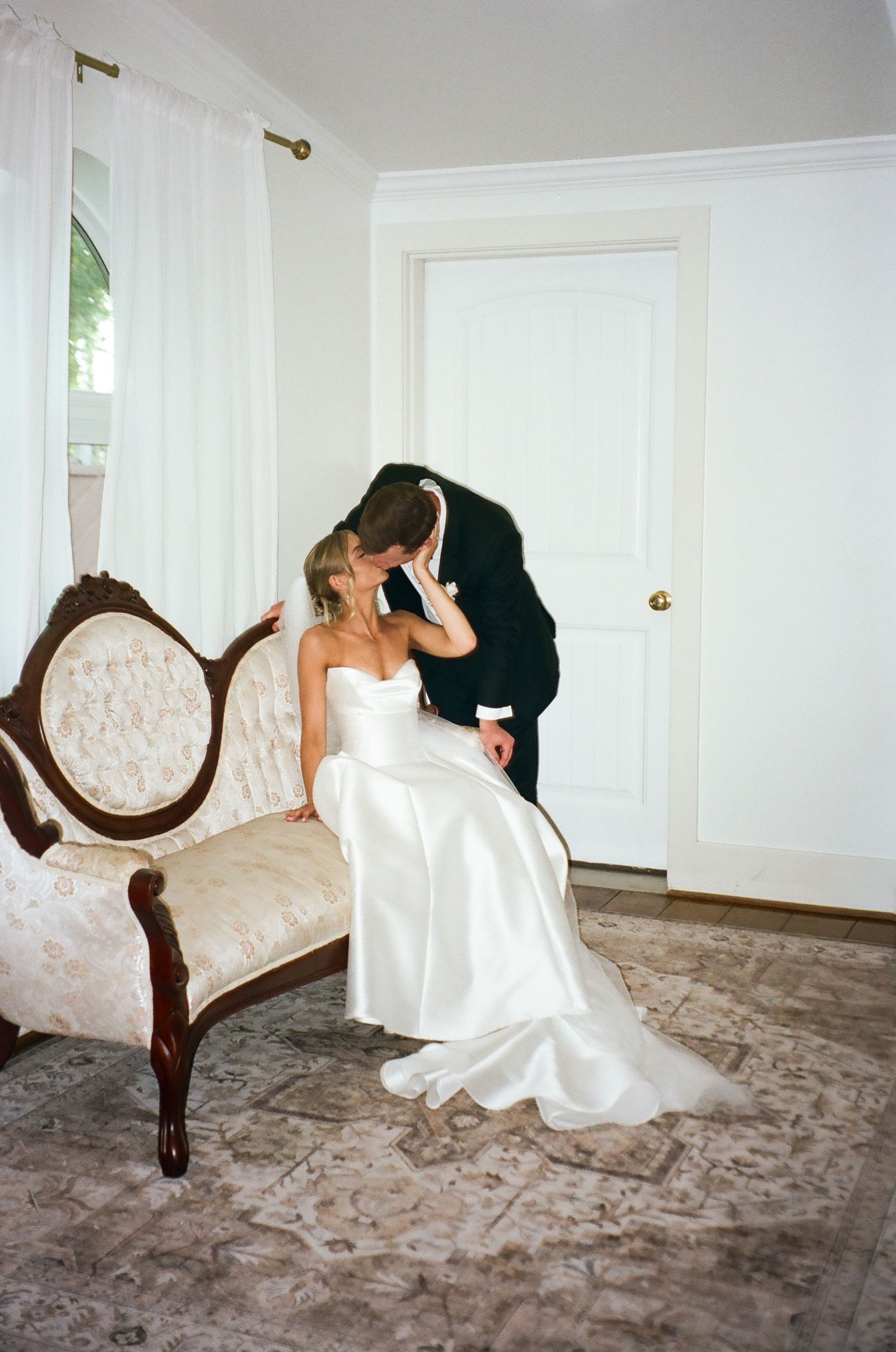 Bride in strapless satin gown seated on vintage settee shares kiss with groom in black tuxedo.