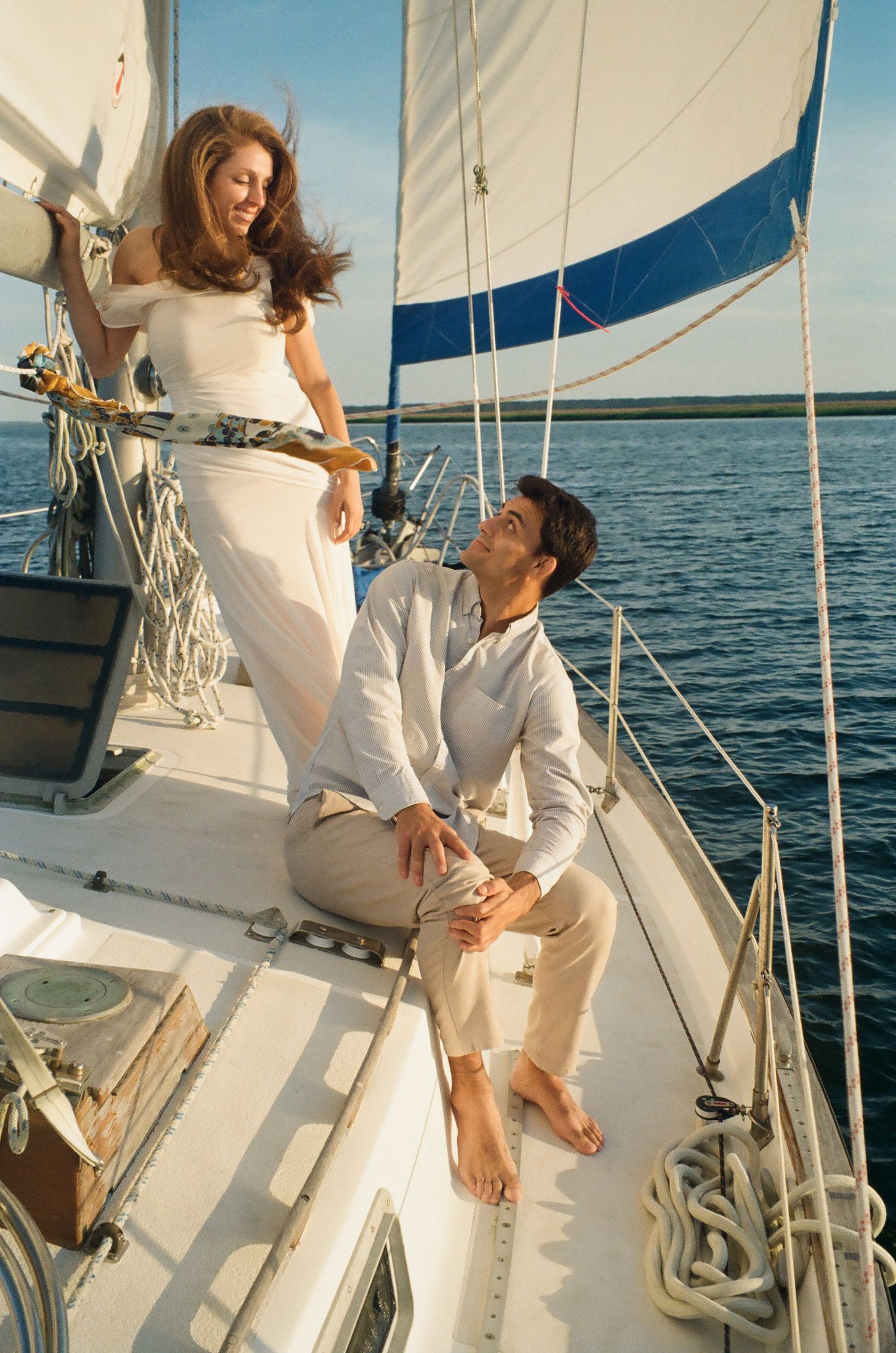 A couple relaxes on a sailboat, woman in white dress standing while man sits gazing up at her on sunny day.