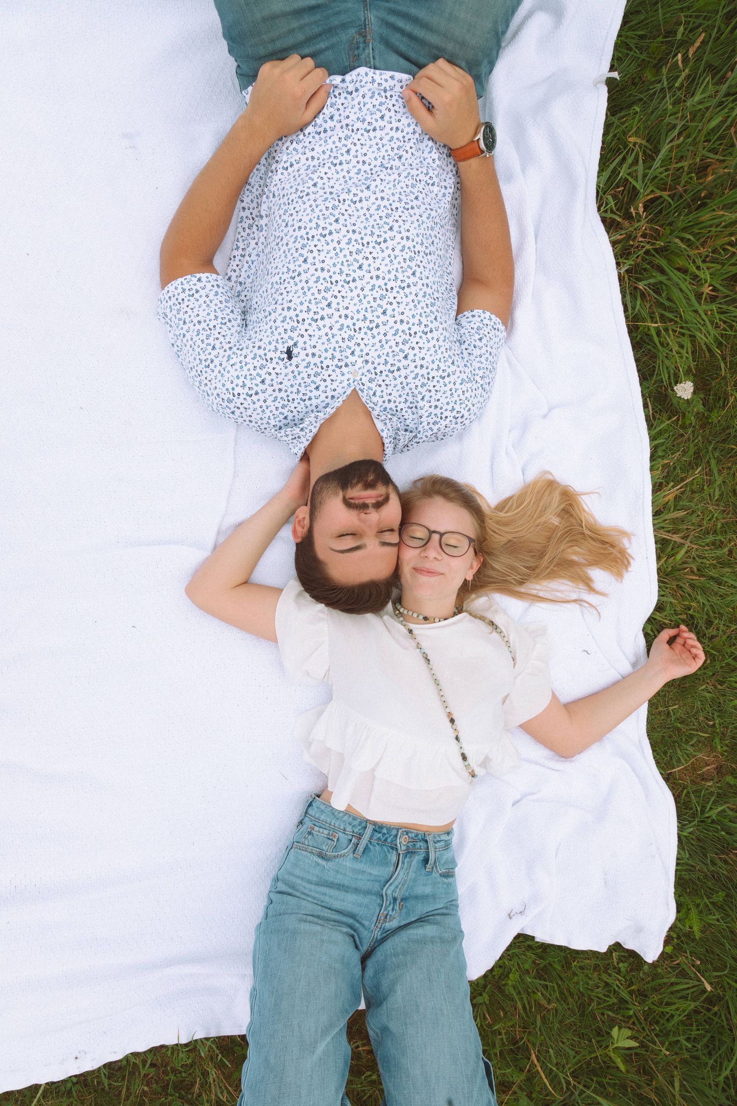 Aerial view of couple lying on white blanket on grass, woman in jeans and glasses smiling.