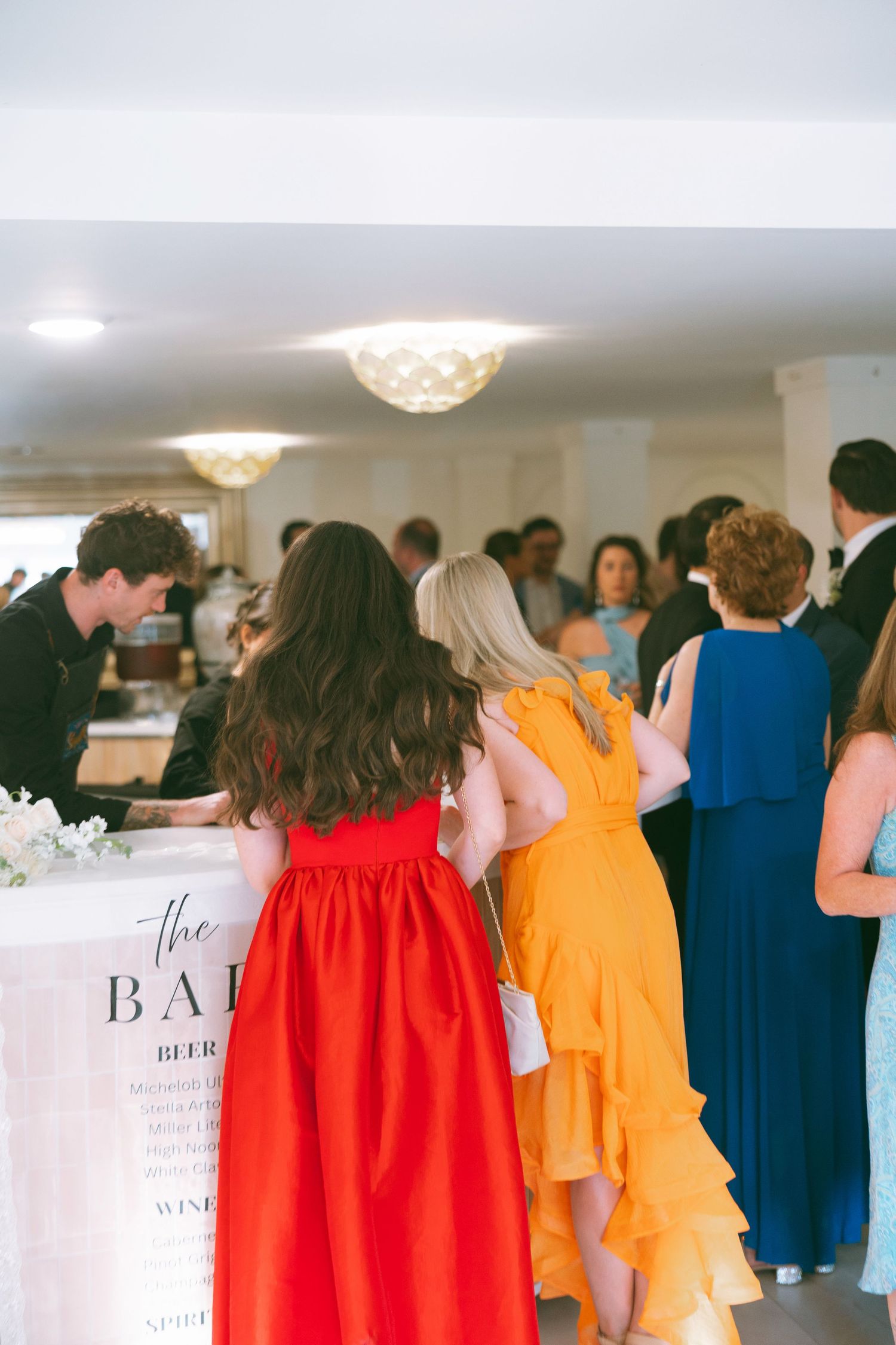 Wedding guests in colorful formal dresses gathered at a bar during a reception.