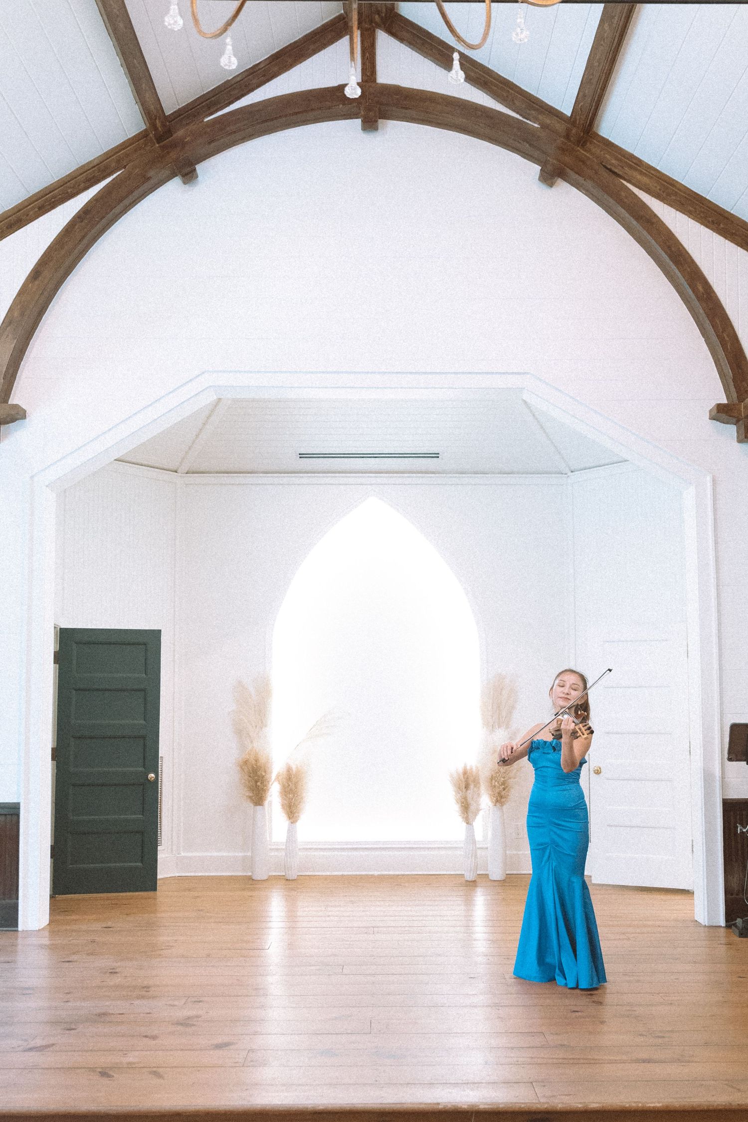 A violinist in a blue gown performs in an elegant white chapel with arched wooden beams and hardwood floors.