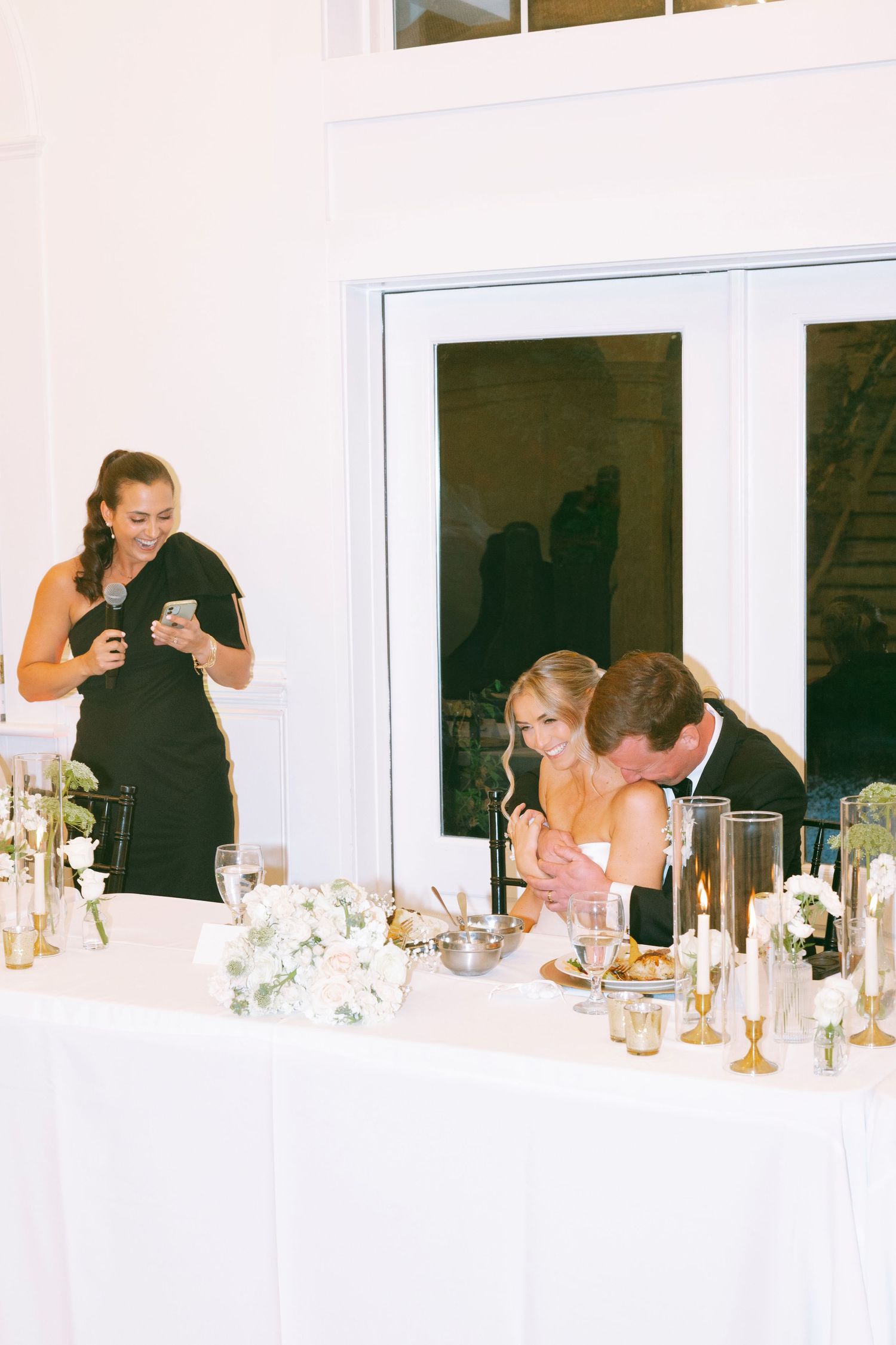 Bride and groom share a laugh at wedding reception head table with floral arrangements and candles.