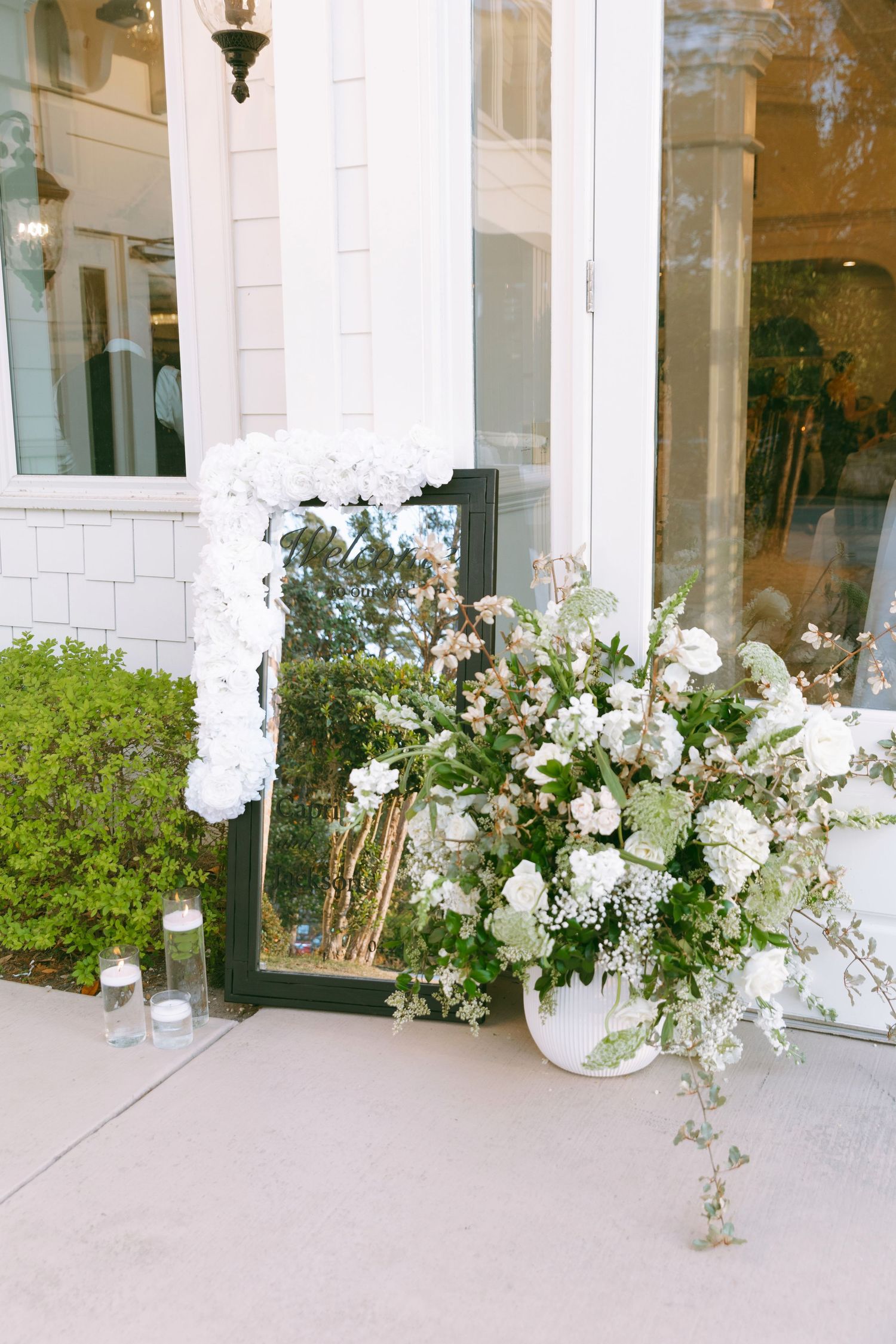 White floral arrangement in large pot beside a mirror leaning against a white building entrance with candles.