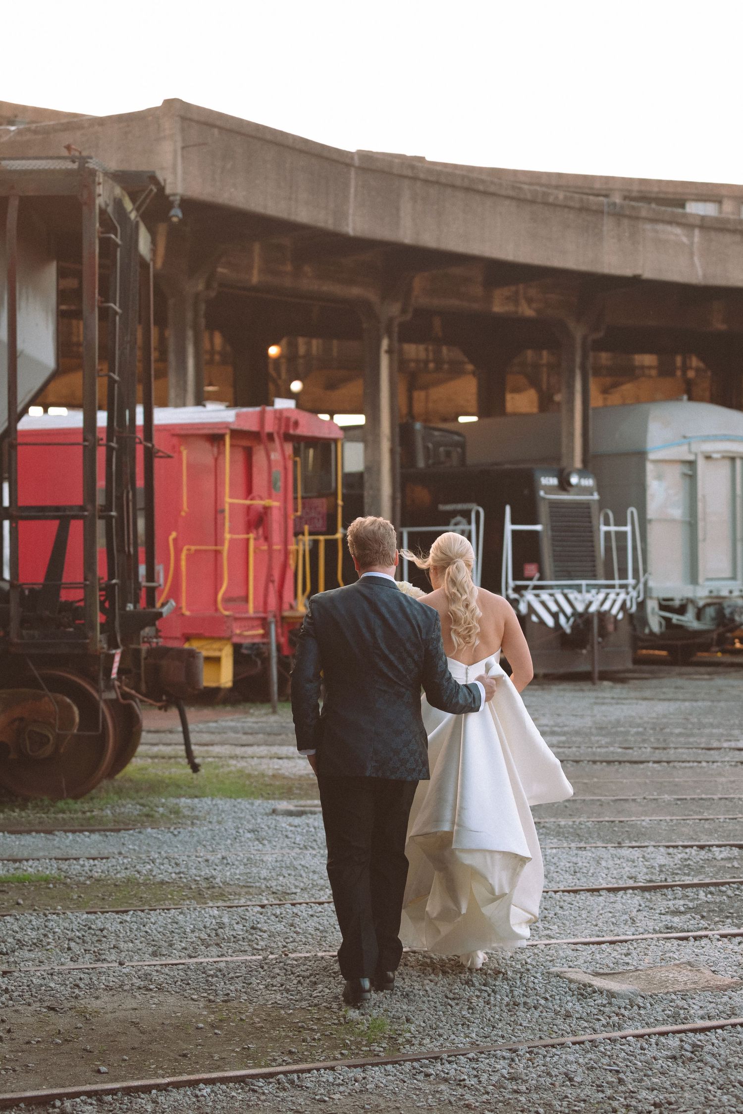 Bride and groom walk hand in hand through vintage train yard during wedding portraits.