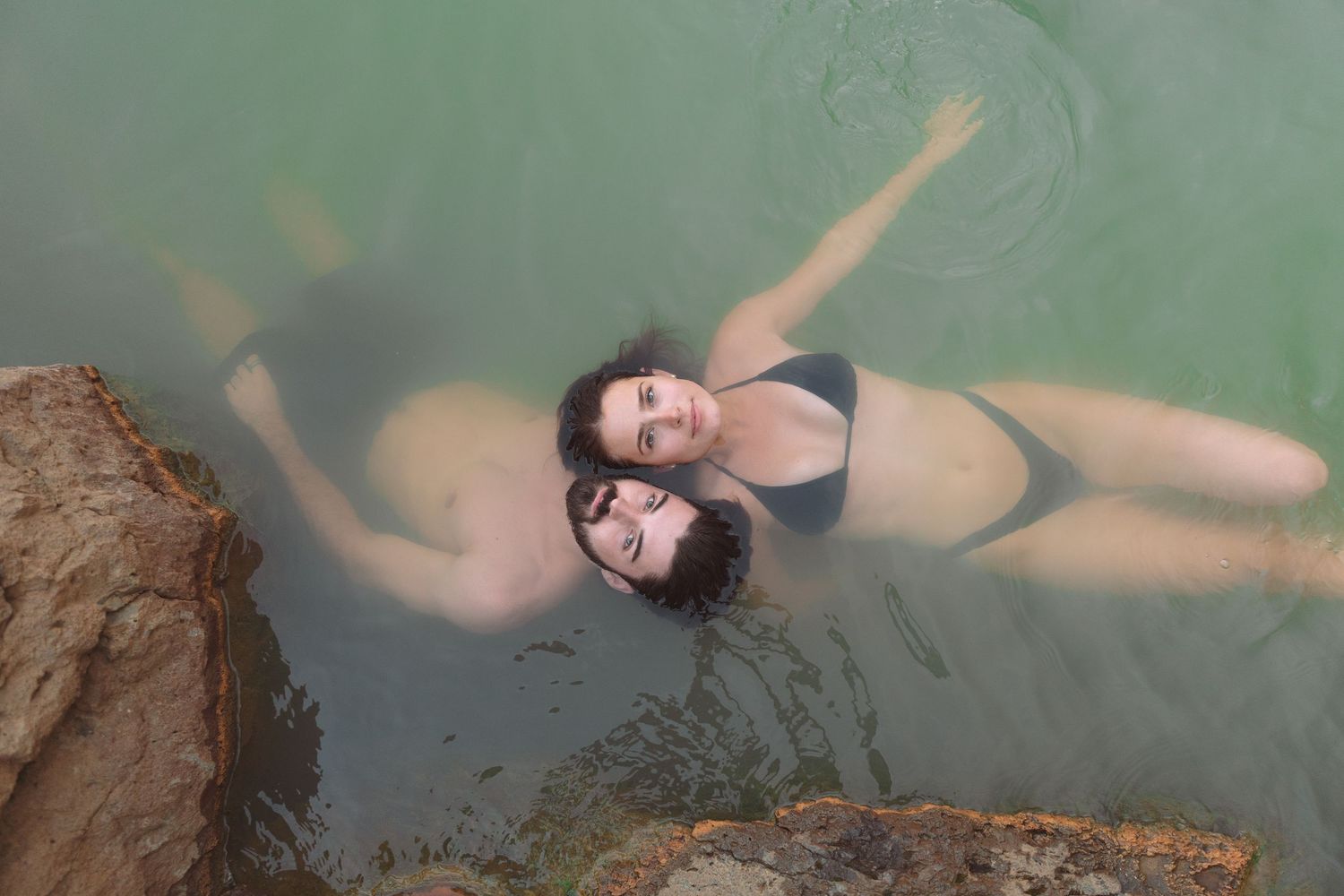Aerial view of couple floating together in a turquoise geothermal hot spring in Iceland.