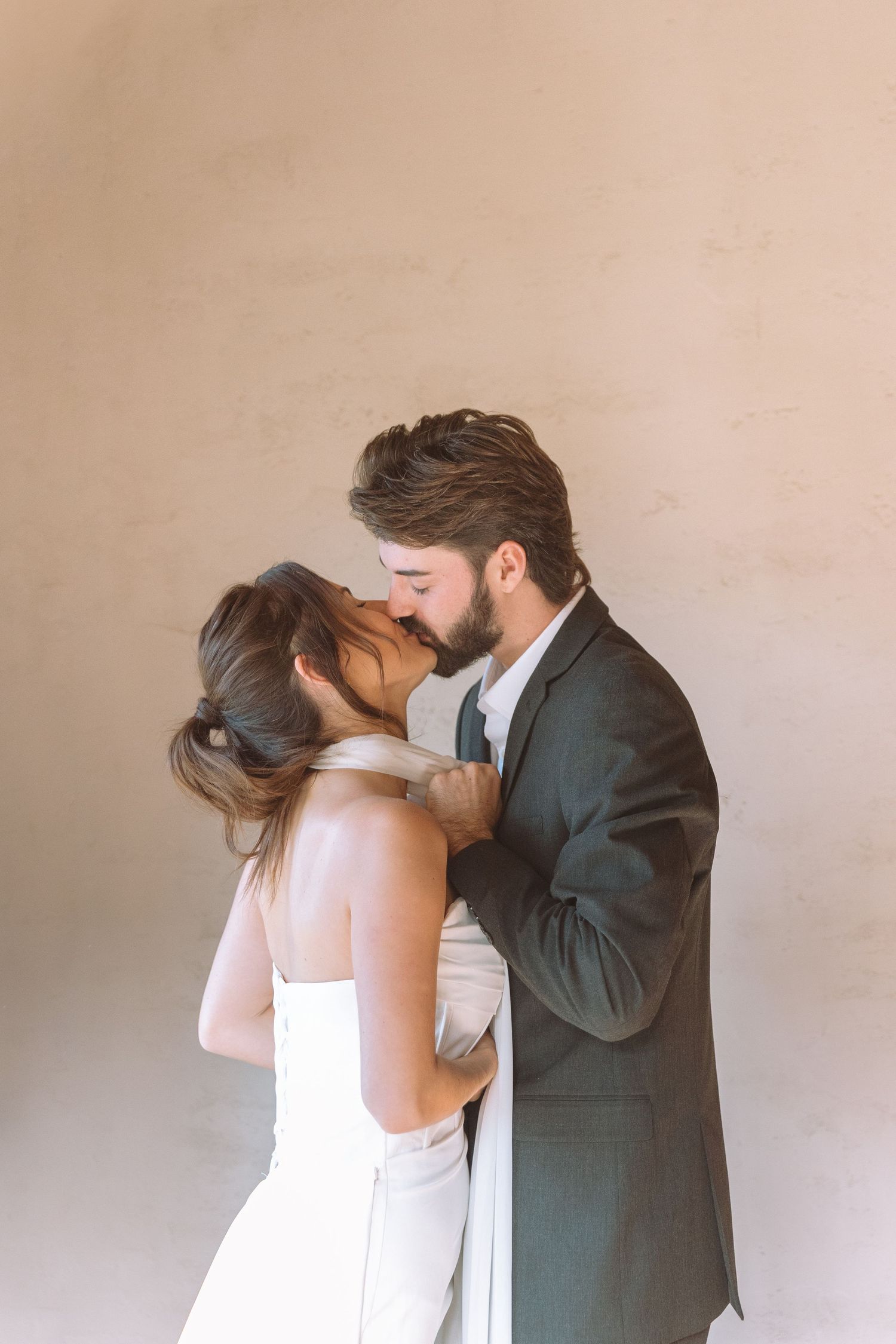 Bride and groom sharing a passionate kiss against a warm neutral plaster wall.