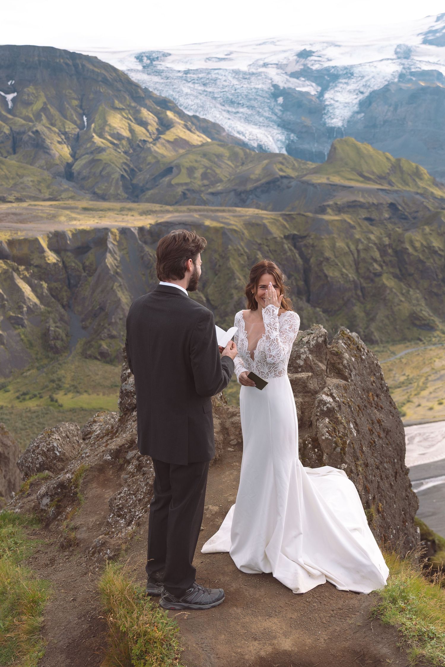 Emotional bride wipes tear as groom reads vows on Iceland cliffside with glacier backdrop.