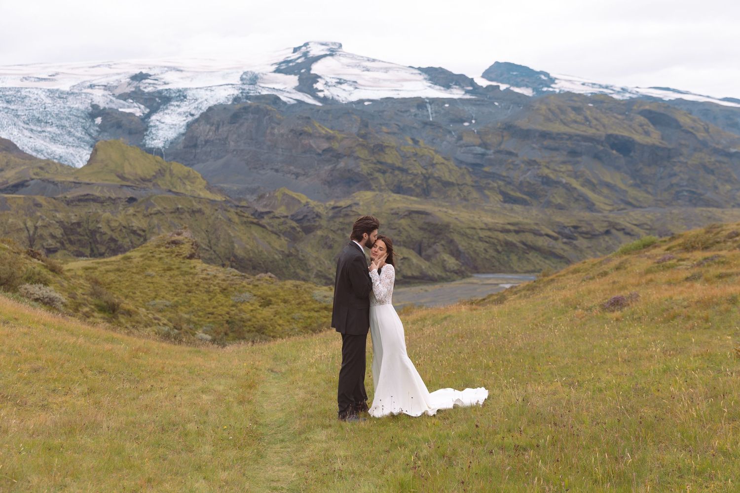 Bride and groom sharing tender moment on green Icelandic hillside with glacier and dramatic mountain scenery.