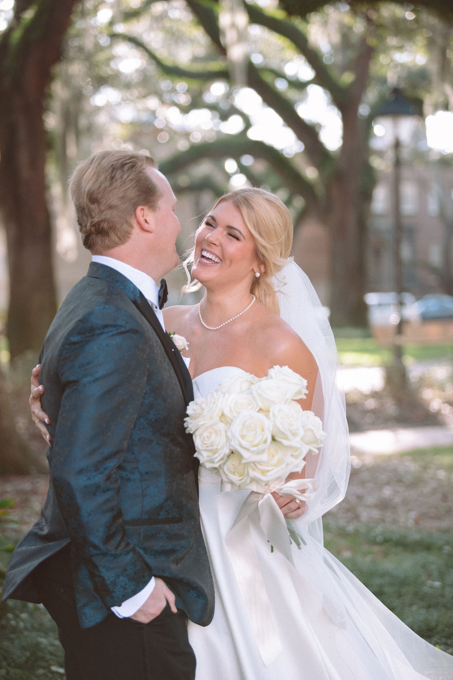 Laughing bride holding white rose bouquet with groom under oak trees in Savannah.