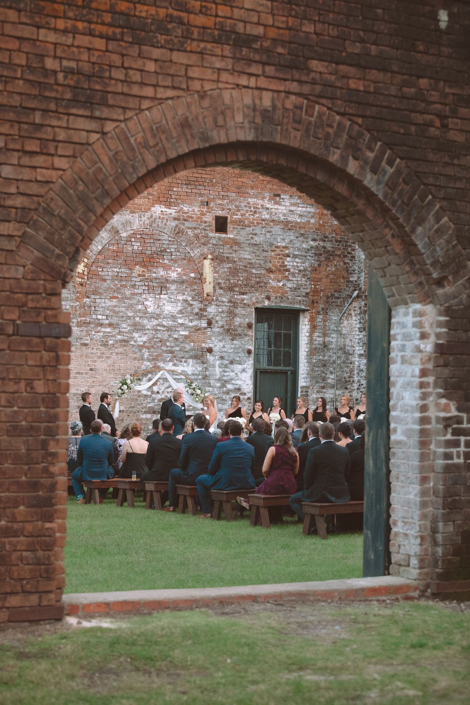 Wedding ceremony viewed through a rustic brick archway with guests seated on wooden benches on green grass.