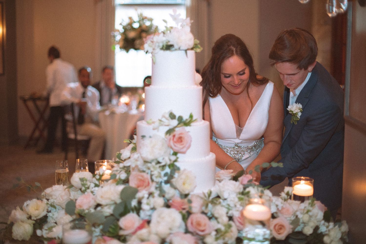 Bride and groom prepare to cut elegant white tiered wedding cake surrounded by pink floral arrangements.