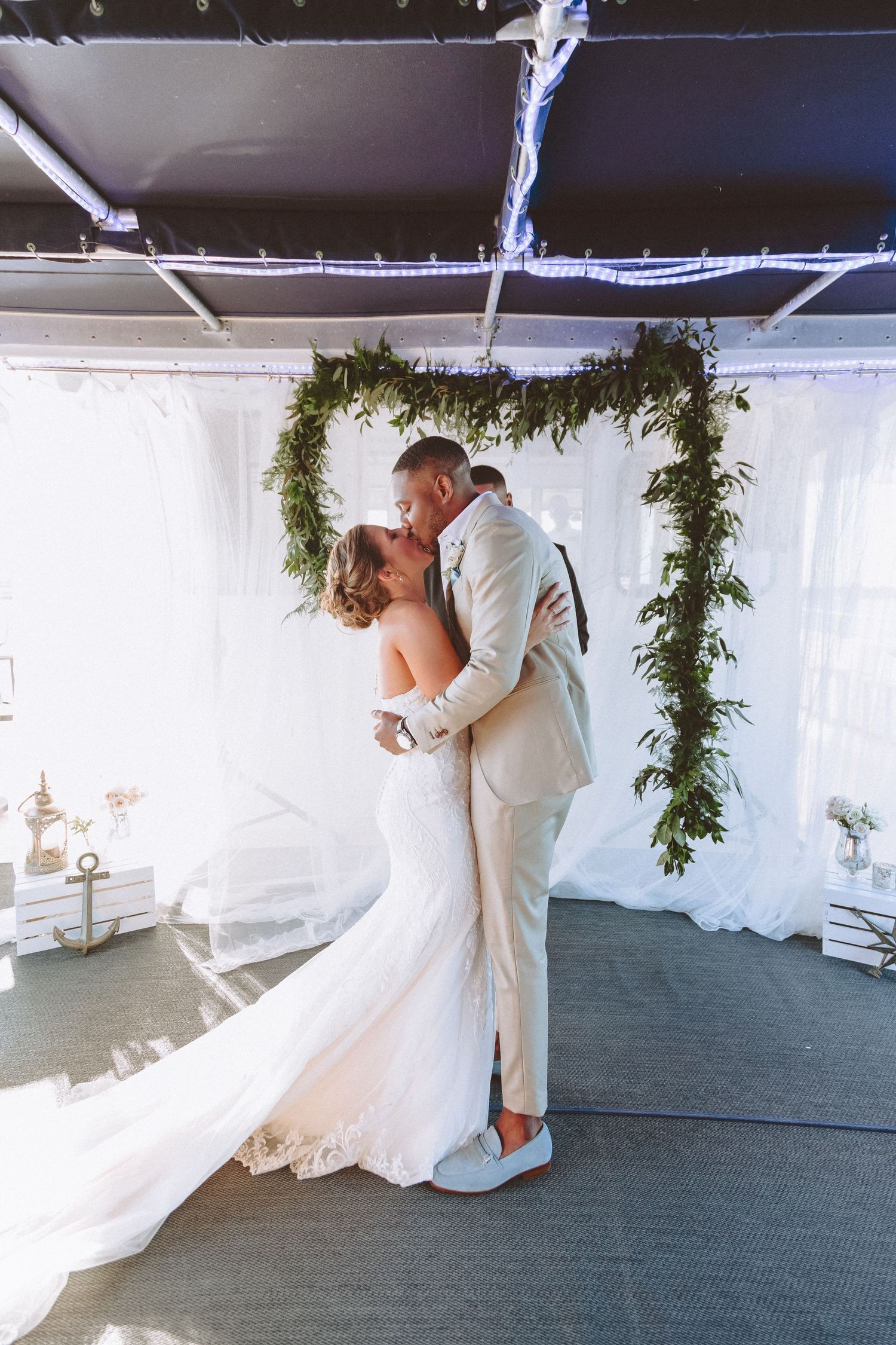 Bride and groom share romantic first kiss under elegant greenery arch at wedding ceremony.