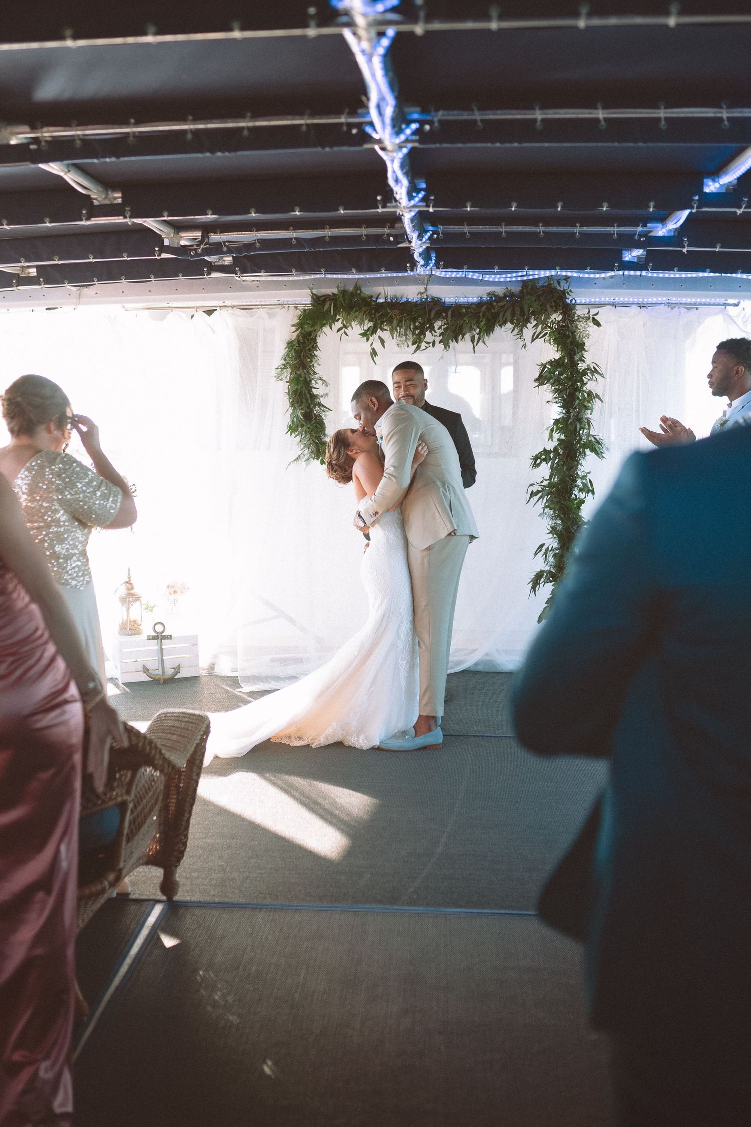 Bride and groom sharing first kiss under a lush greenery arch at indoor wedding ceremony.